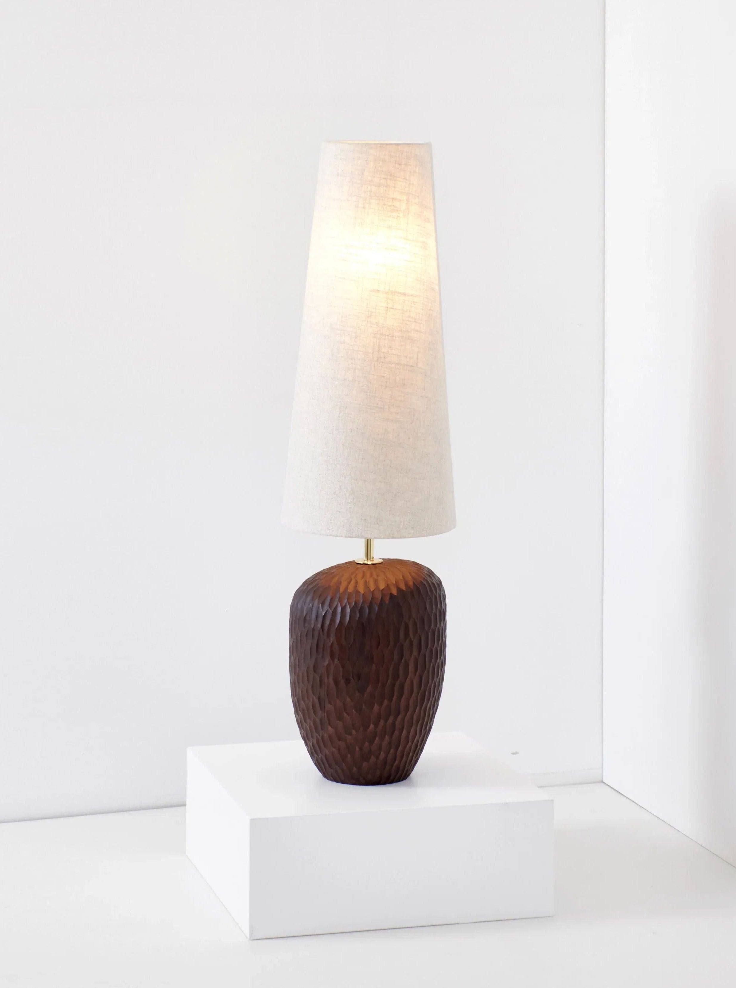 A Large Foz Lamp by Project 213A with a textured, solid wood base and a tall, tapered white lampshade is displayed on a white pedestal. The lamp is set against a minimalist white background.