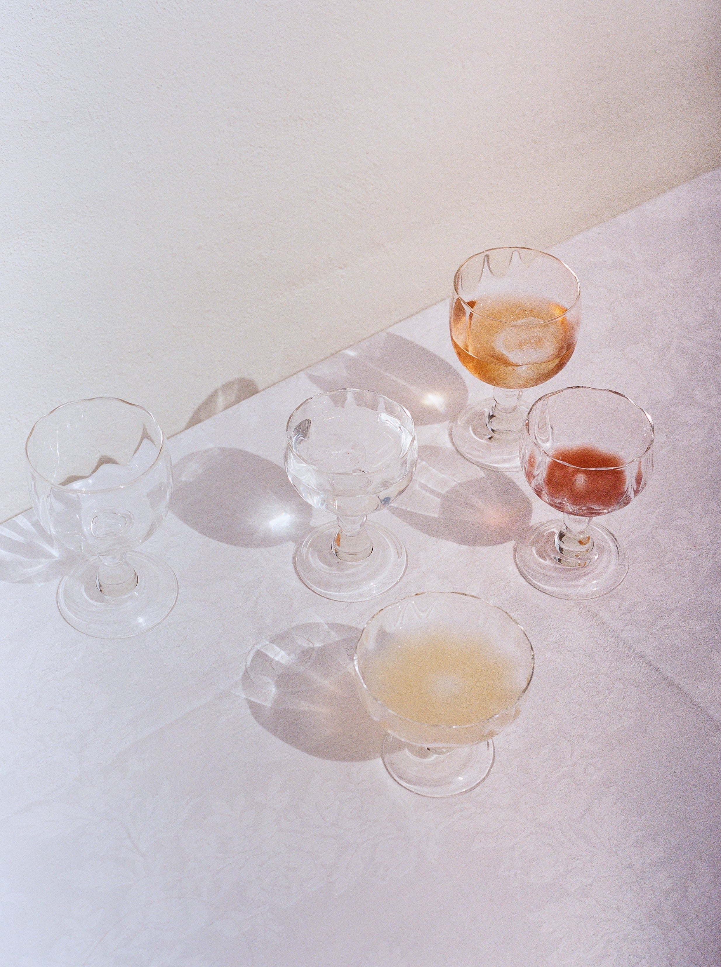 Five vintage Akua Objects Alban Champagne Glasses on a lace tablecloth, with varying levels of a pinkish beverage, illuminated by soft sunlight.