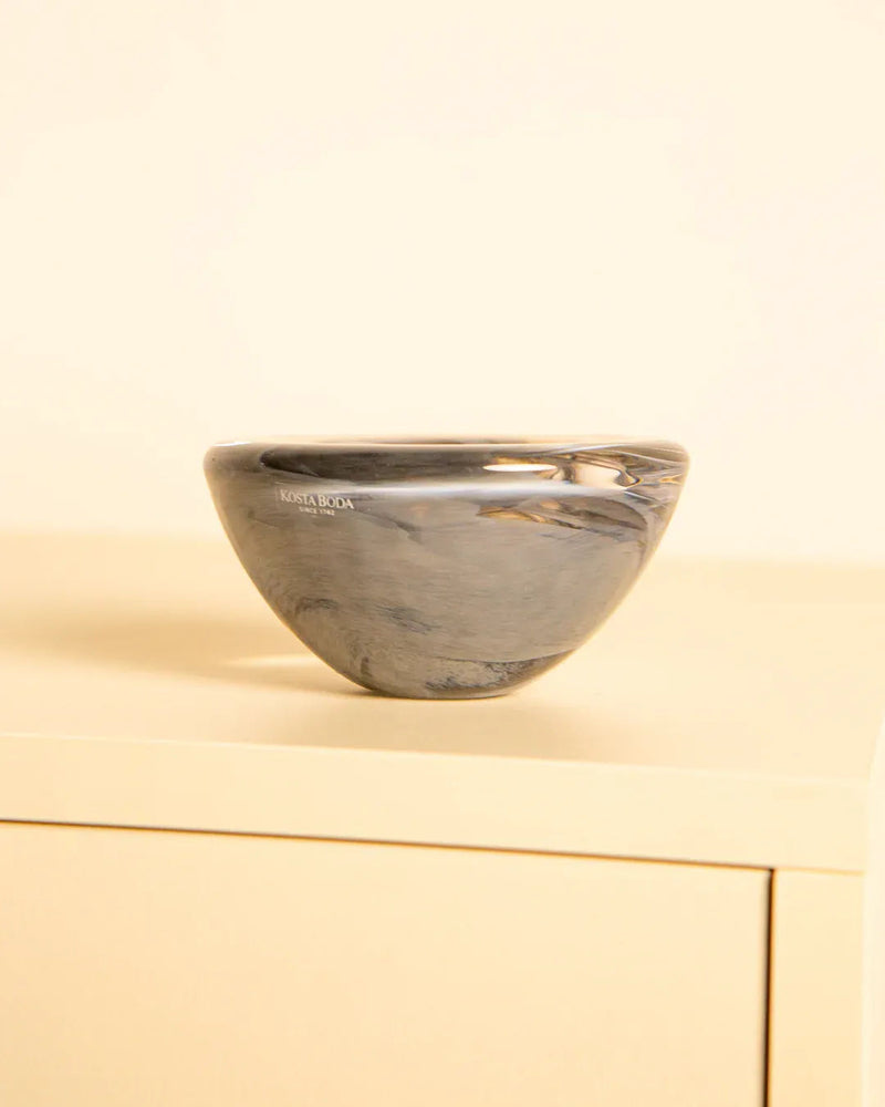 The Vide-poches "Fumé" by Kosta Boda 80's from Treaptyque—a small, round, polished gray marble bowl—rests on a light beige cabinet against a pale background, showcasing Scandinavian elegance.