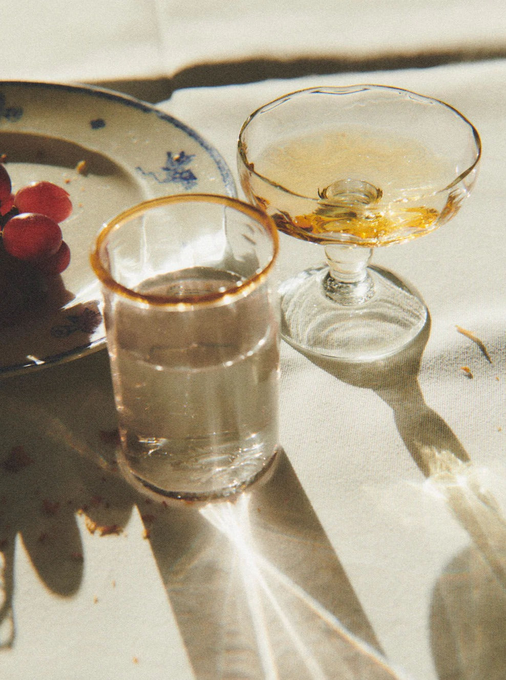 A still life photo featuring a glass of clear liquid and an Akua Objects Alban Champagne Glass - Set of Four with amber liquid, beside a plate with grapes. The scene is bathed in dappled sunlight, casting soft shadows on.