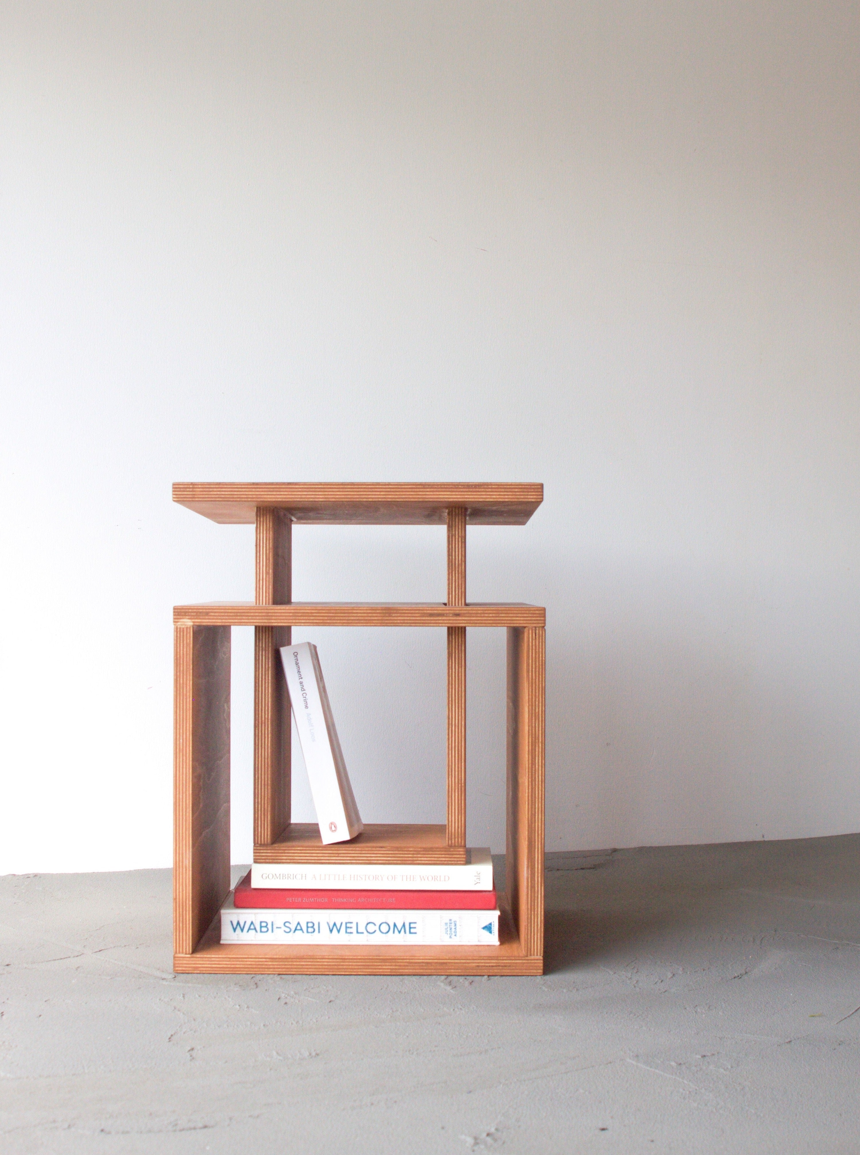 A minimalist birch plywood side table from Edoardo Lietti Studio with three levels, featuring two books placed horizontally on the bottom shelf, positioned against a plain light-colored wall.