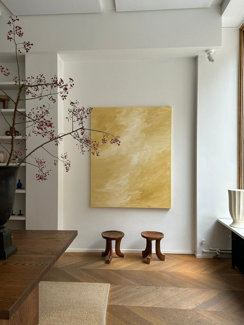 A minimalist room features "Sands of Light," a large mixed media canvas by Mattias Storm, on a white wall, with two small wooden stools, a tall vase of red berry branches, and natural light streaming onto a chevron wood floor.
