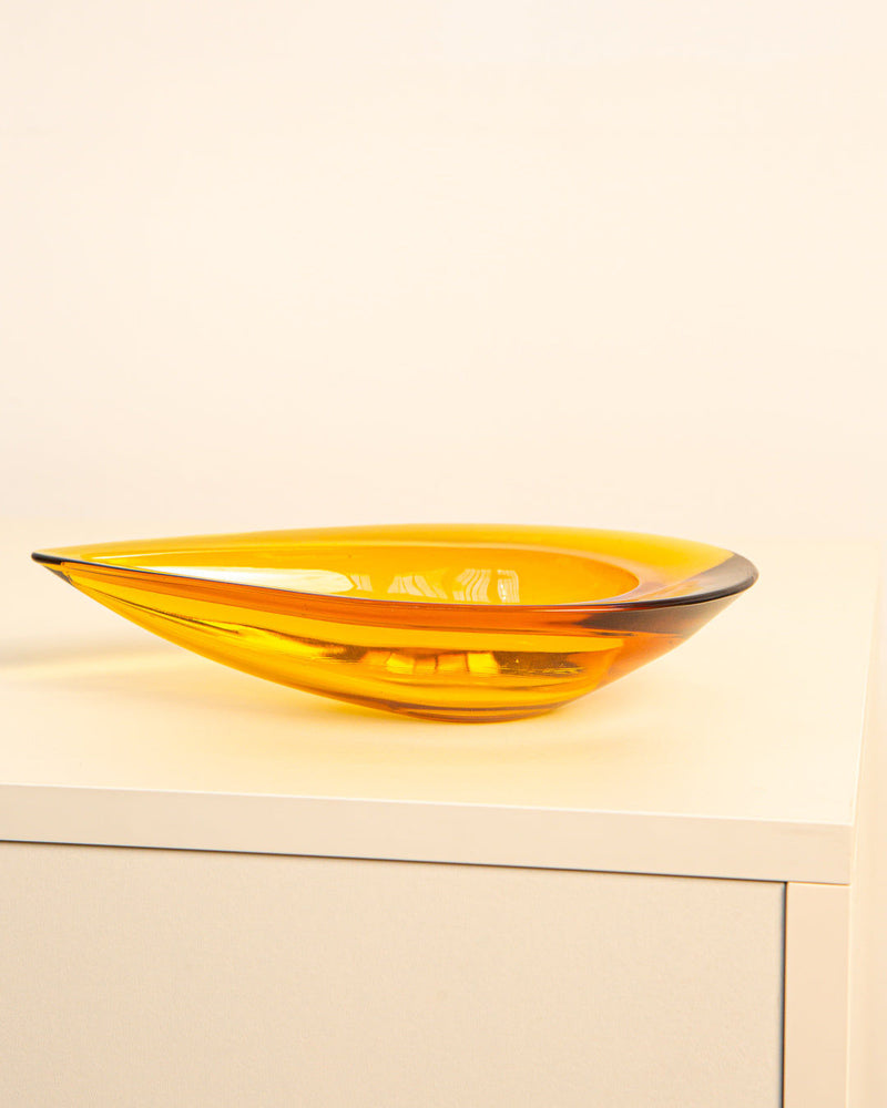 The Treaptyque Large Italian Vide-poches "Poire" 60's, a modern amber glass bowl with a sleek teardrop form inspired by 1960s design, is displayed on a white cabinet against a light beige background.
