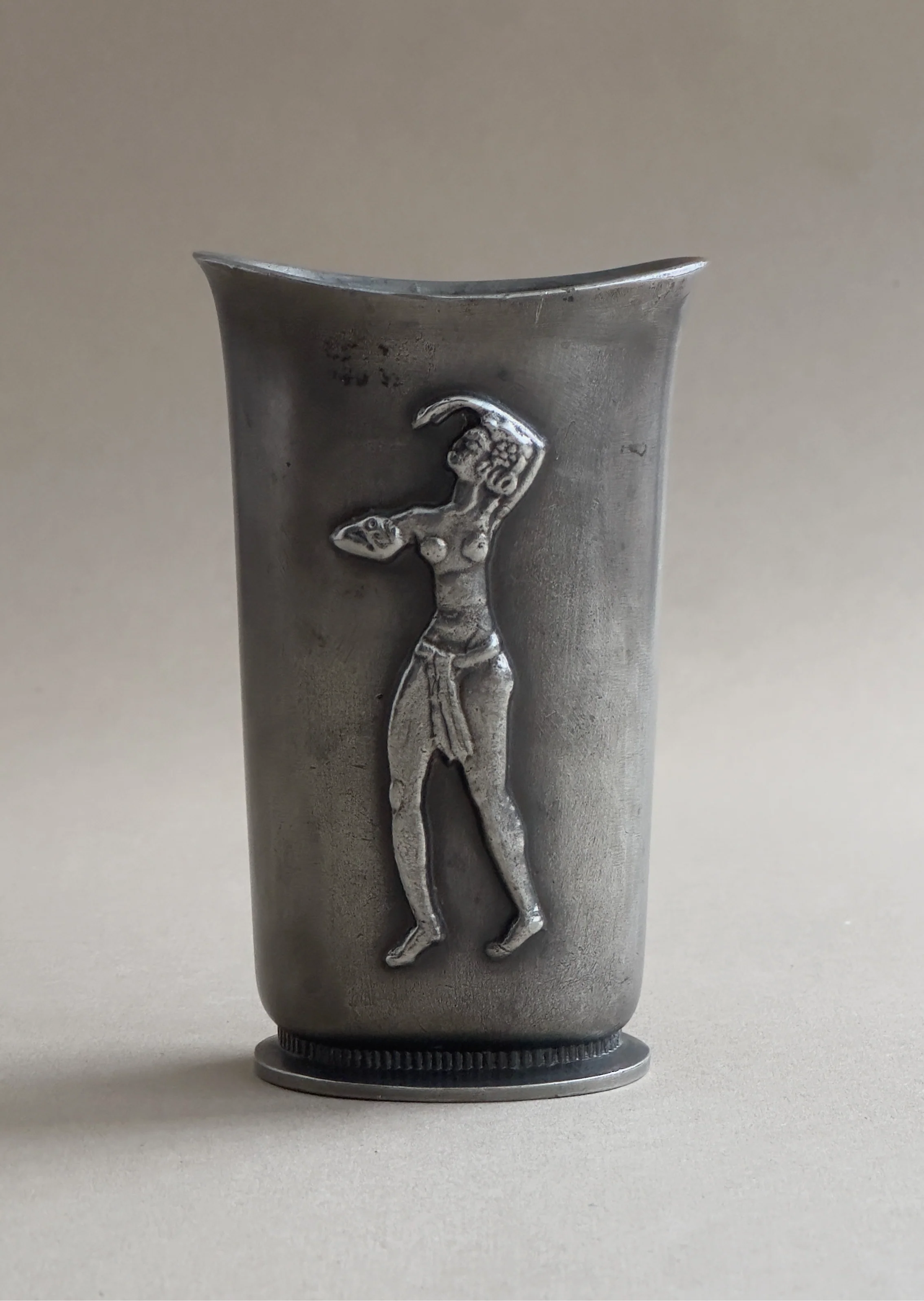 The Art Deco vase in cast pewter by Galerie Storm, reminiscent of a Swedish Grace vase, features a raised relief of a dancing figure in a dynamic pose. Its plain, neutral background enhances the elegance of the design and highlights its captivating pewter finish.