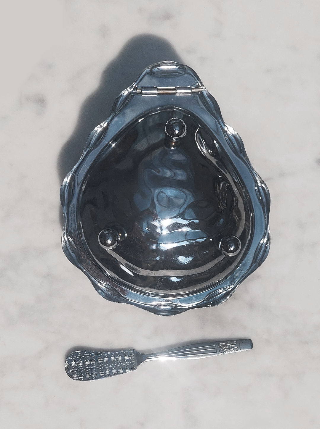 A small, shiny Shell Butter Dish from Les Objoies rests on a light marble surface. Below it lies a silver-toned, ornate spoon with a textured handle. The butter dish appears to be closed, reflecting light off its polished surface.