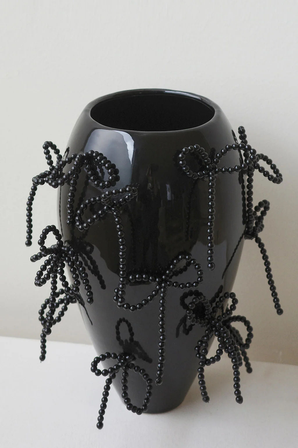 The Pearly Pearl by Completedworks is a large, handcrafted ceramic vase in gloss black, featuring multiple black beaded bows that add texture and decoration. It's set against a plain white background for a sleek, modern display.