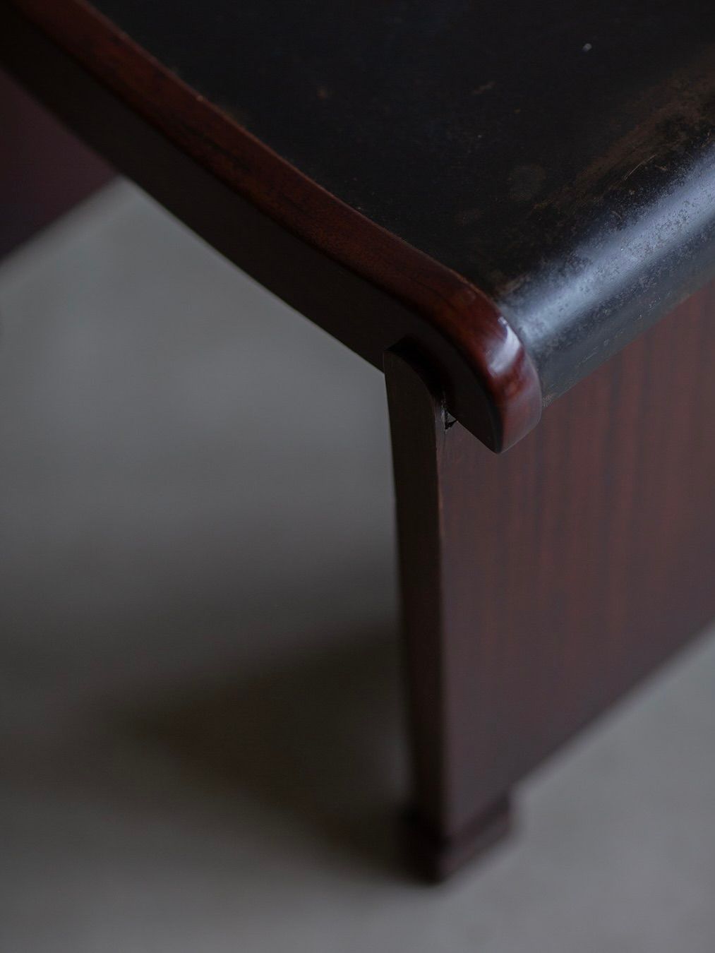 Close-up of a dark wooden corner from the Bicci de' Medici Pierre Chareau Curule Stool (Model MT 1015), showcasing an Art Deco elegance with a smooth, shiny finish. It features an aged appearance with visible grain and subtly curved edges against a muted background.