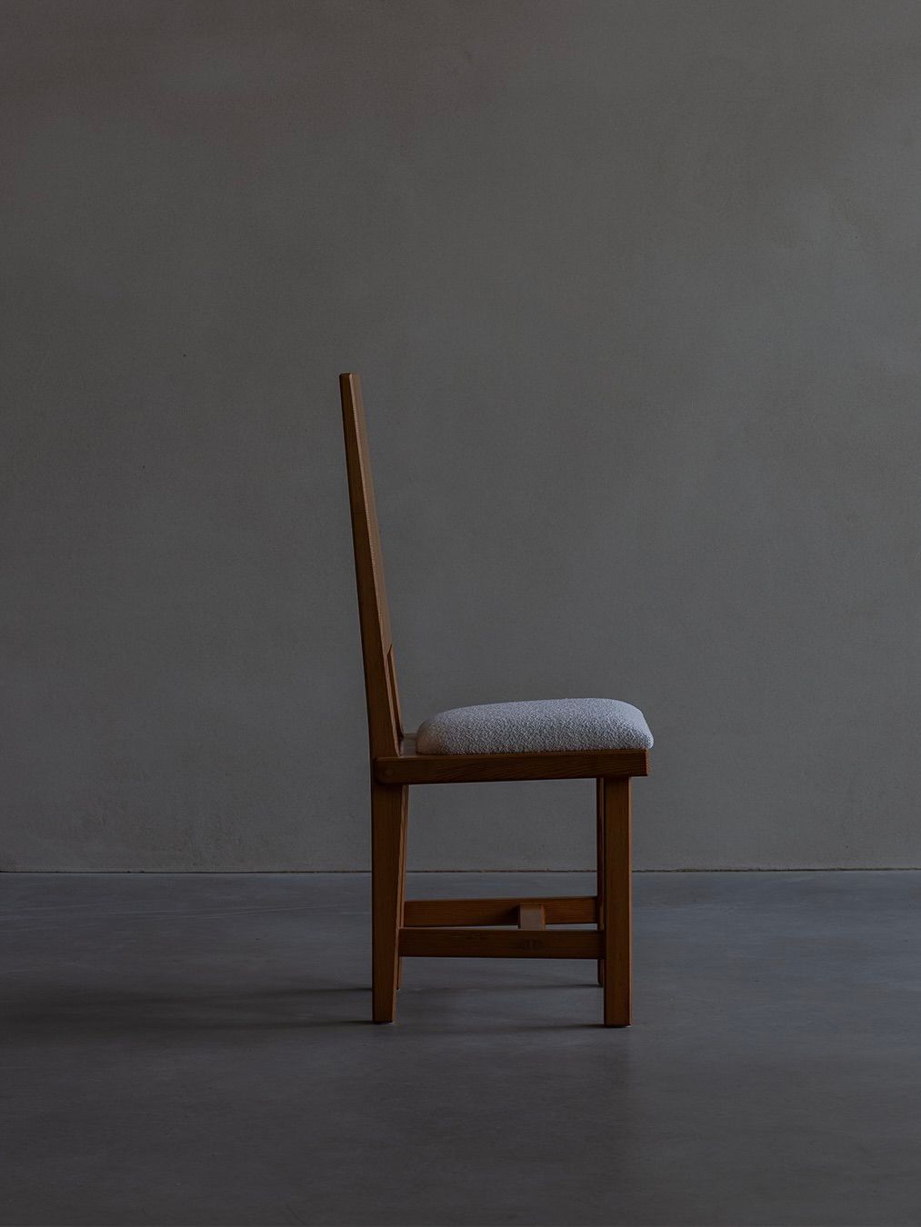 In a profile view against a dimly lit gray background, the Bicci de' Medici Göran Malmvall Model 553 wooden chair features a straight backrest and cushioned seat, highlighting its simple and contemporary Swedish design aesthetic.