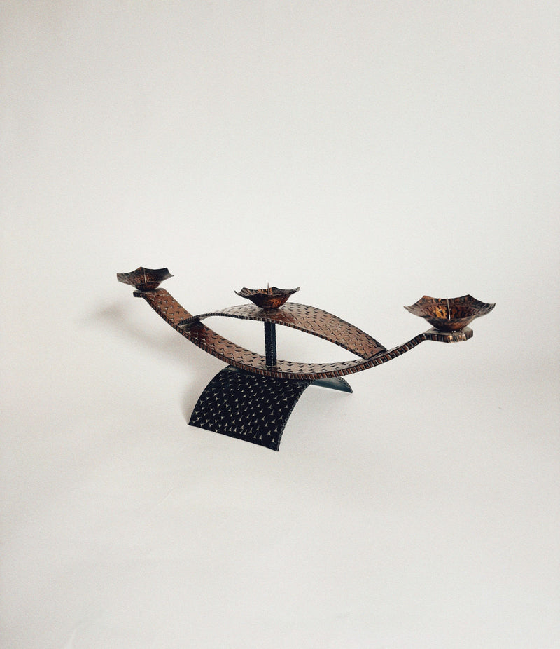 The Huerto Two Tone Metal Candleholders feature a sculptural, handmade design with a curved, textured black metal base and three leaf-shaped holders, displayed against a plain white background.