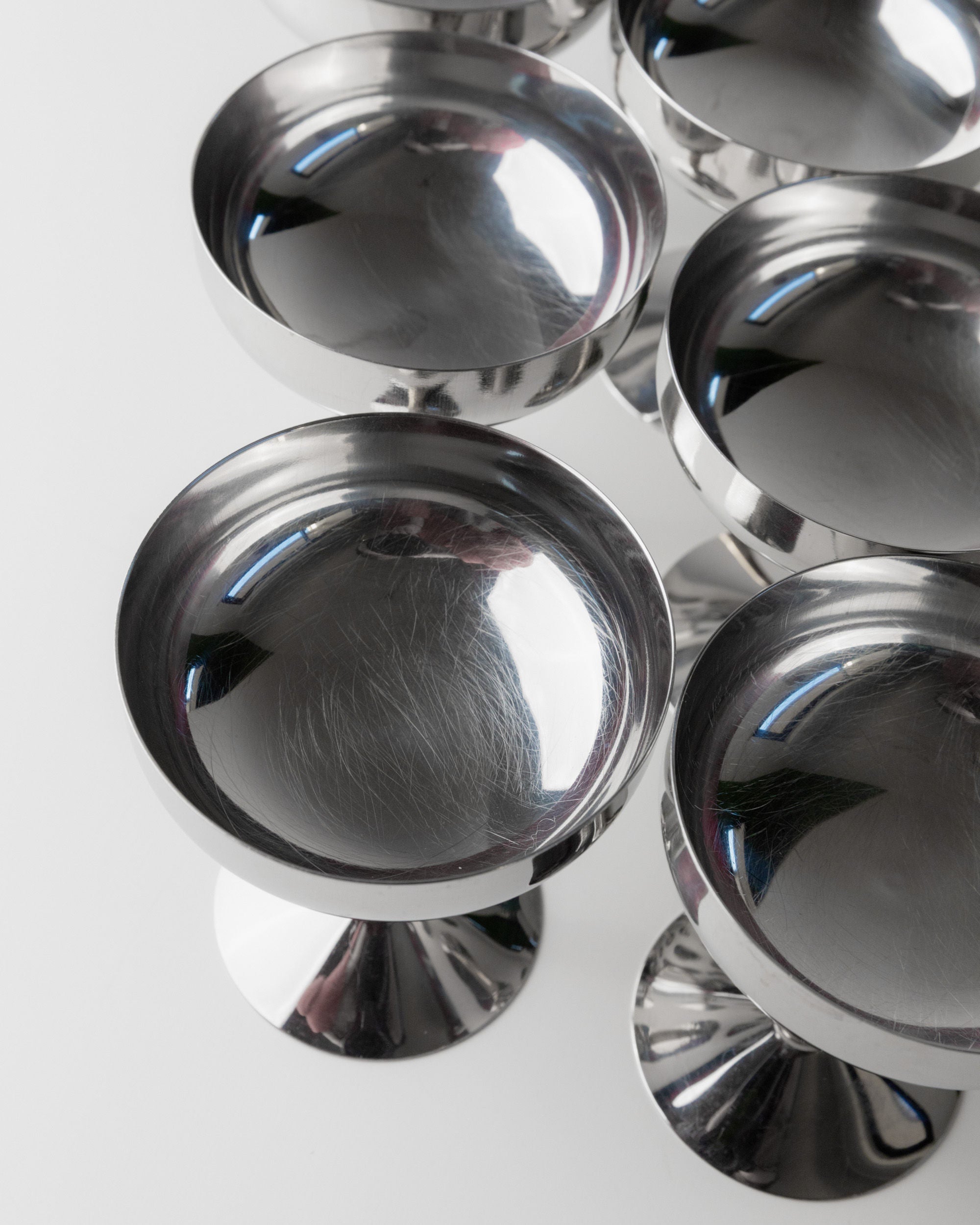 Close-up of several polished Bottega Jacobs vintage stainless steel champagne coupes from the '70s, with round bases, arranged in rows. The smooth, reflective surfaces capture highlights and reflections against a plain white background.