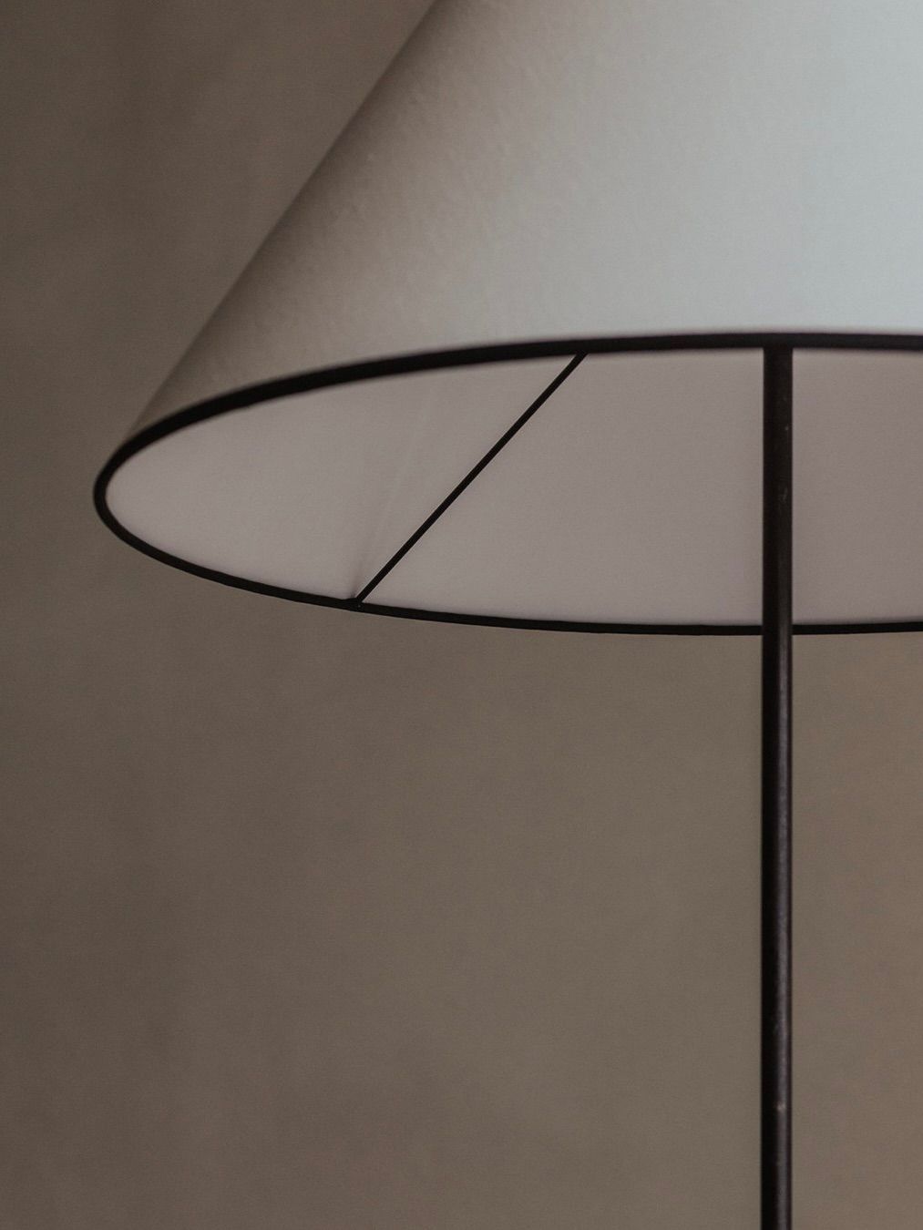 A close-up features the "Manner Of Royere & Adnet - Wrought Iron Tripod Lamp" by Bicci de' Medici, showcasing its white, tapered lampshade and sleek black metal stand against a softly lit neutral background. The minimalist design echoes mid-20th-century aesthetics with clean lines and simplicity.