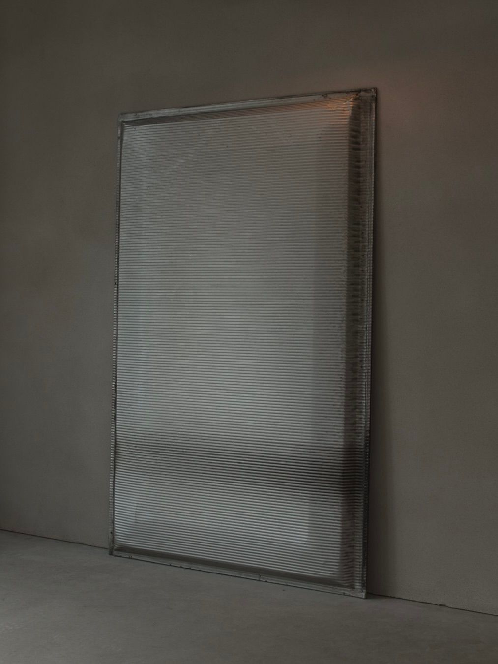 Bicci de' Medici's Jean Prouvé - Façade Panel 108, with its rippled metallic surface, leans against a plain dark gray wall. The texture casts a subtle interplay of light and shadow, exuding a modern minimalist aesthetic.