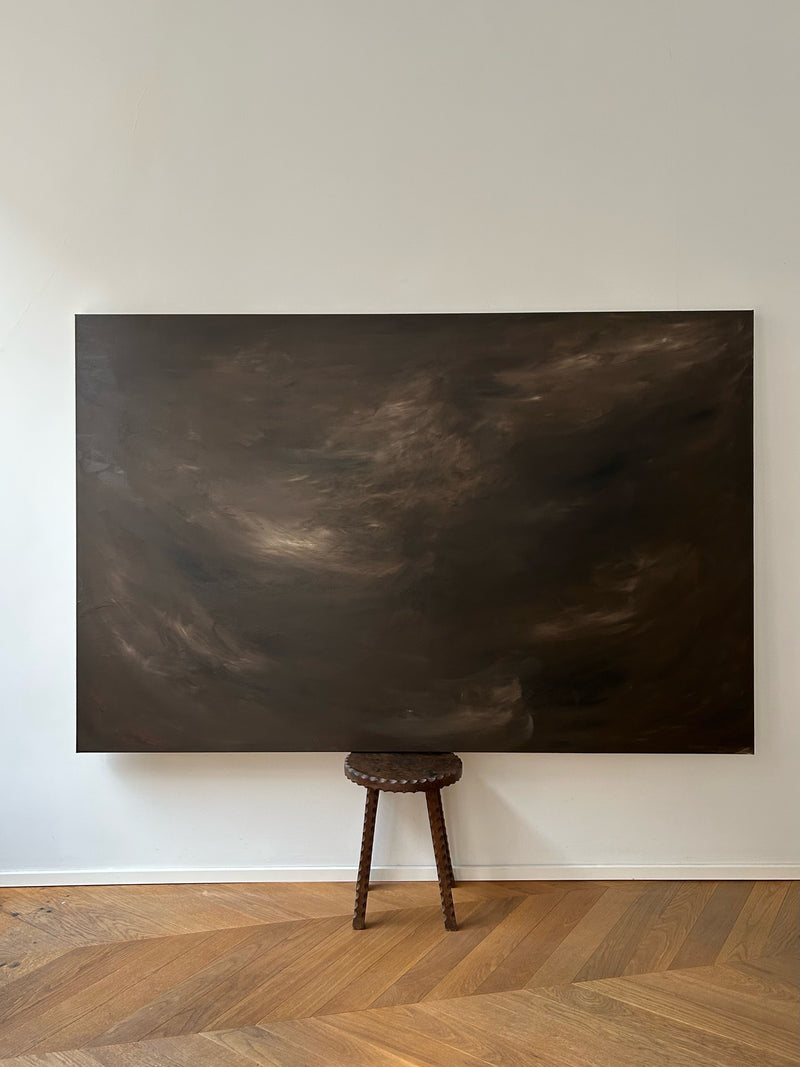 The "Veil of Shadows" painting by Mattias Storm, a large abstract work featuring swirling, cloud-like textures in mixed media on canvas, hangs on a light-colored wall. Below it stands a small, round wooden stool with carved legs, perfectly complementing the artwork above as it rests on the parquet floor.
