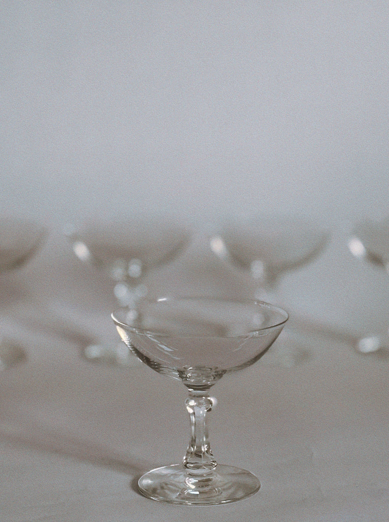 A clear vintage-style coupe glass from the Huerto Set of 5 Cocktail Glasses is centered in focus, while several matching petite cocktail glasses are softly blurred in the background against a neutral surface.