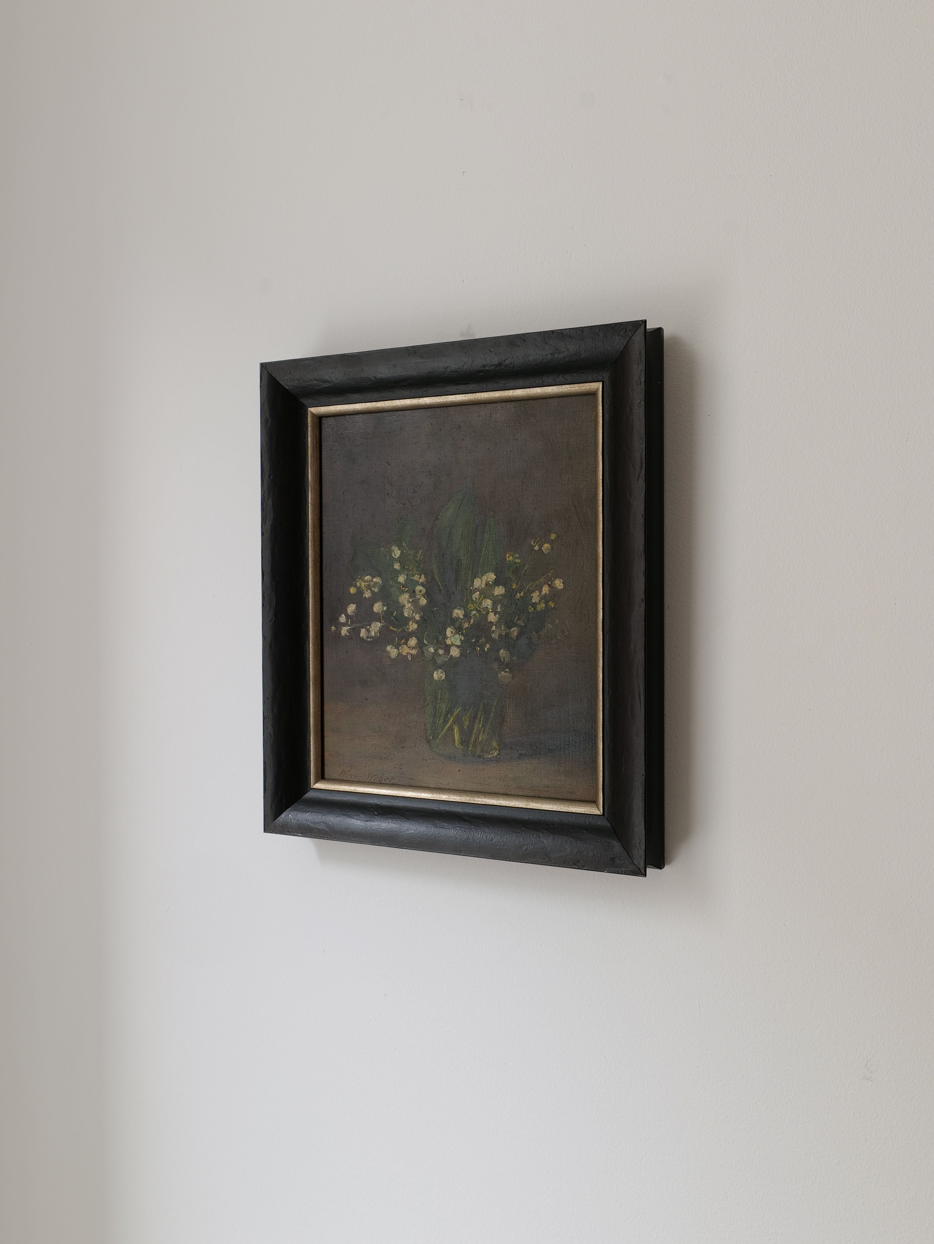 A framed painting of a floral arrangement hangs on a plain light-colored wall. This exquisite Collection apart art piece, "Lily of the Valley Still Life 1961," depicts a bouquet of small white flowers, possibly Lily of the Valley, with green leaves in a vase, set against a dark background. The frame is simple and black, with a slight shine.