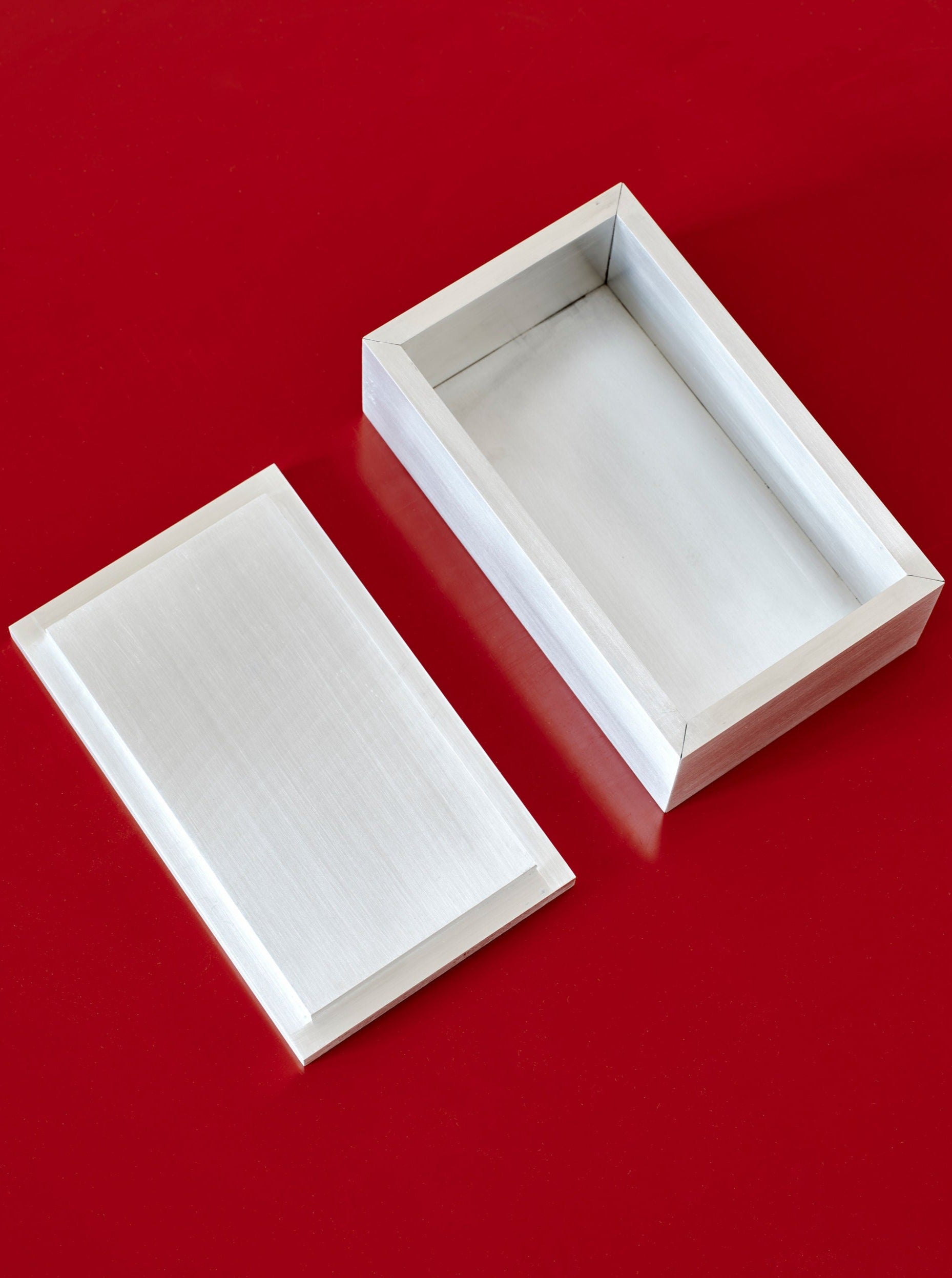 A silver rectangular Miter Box, crafted from high-grade material by MOCK STUDIO, with a detached lid placed next to it, set against a vibrant red background. The brushed metal boasts a smooth, reflective surface, and the box appears to be empty.