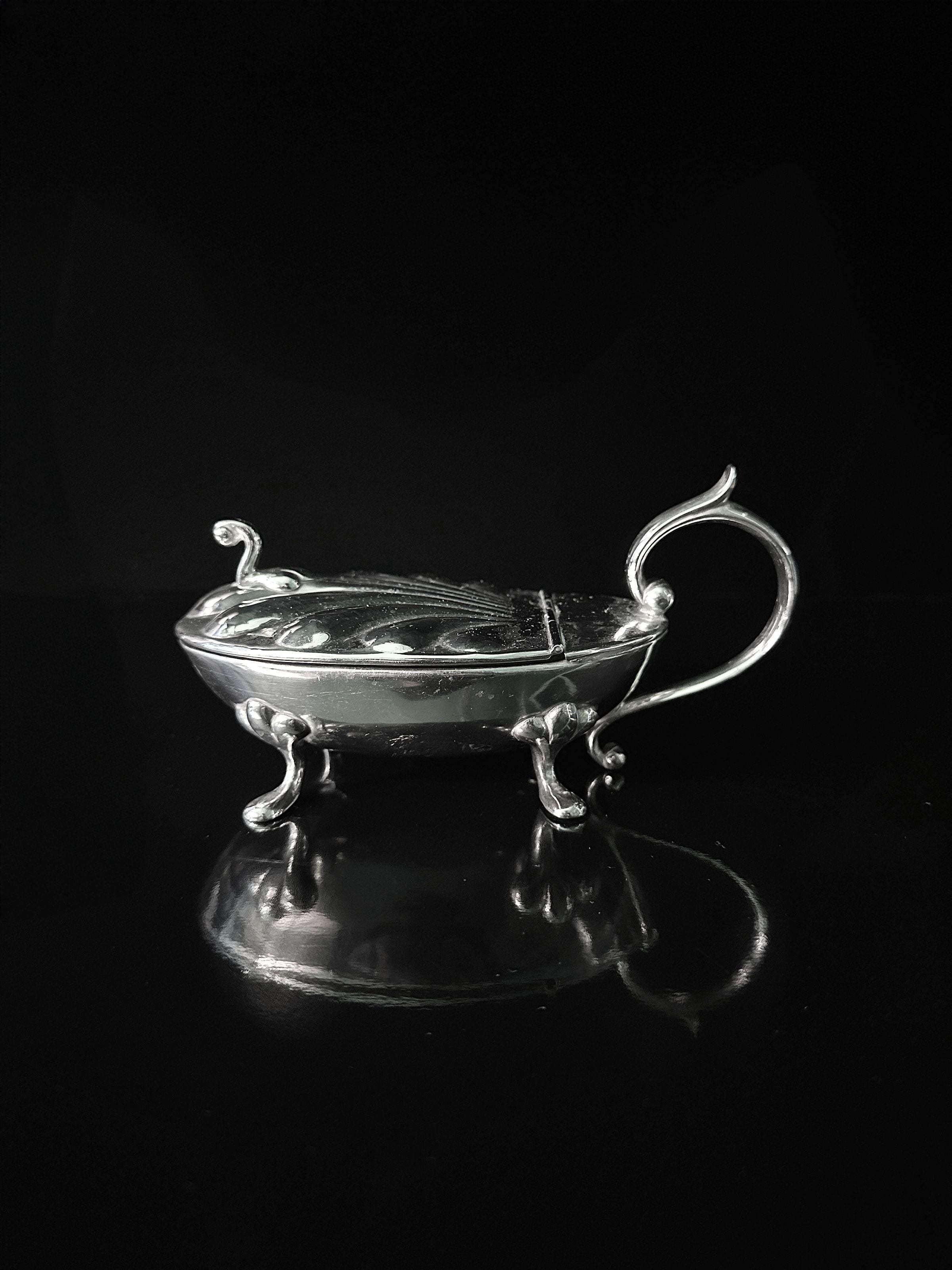 The Silver-plated Caviar Frog 1950s by Dodo Vintage features a reflective, ornate design with a lid and elaborate handle that resembles a refined caviar container. Intricate designs and three elegantly curved legs cast a subtle shadow against a black background.