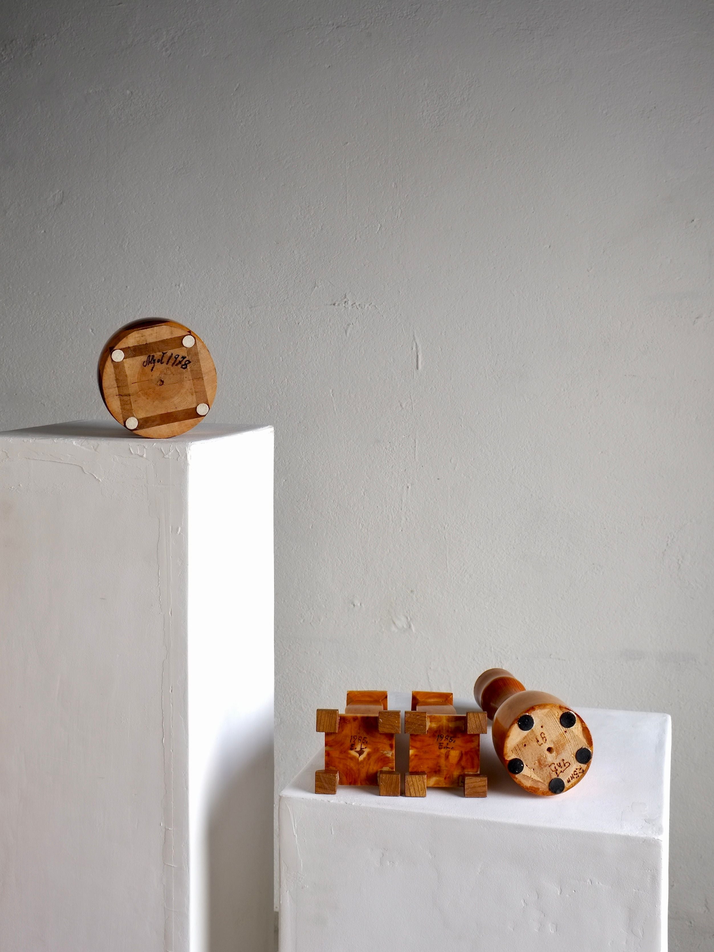 Displayed on two white pedestals against a plain gray wall are three exquisite pieces from the Veter Vintage collection: Carved Wooden Candle Holders and Bowl Set from Sweden, circa 1970s. The lower pedestal showcases a cylindrical candle holder and a block puzzle, while the taller pedestal features an upright round bowl. Each piece highlights distinct wood grain and remarkable craftsmanship reminiscent of vintage carved wooden decor from the 1970s-1980s.