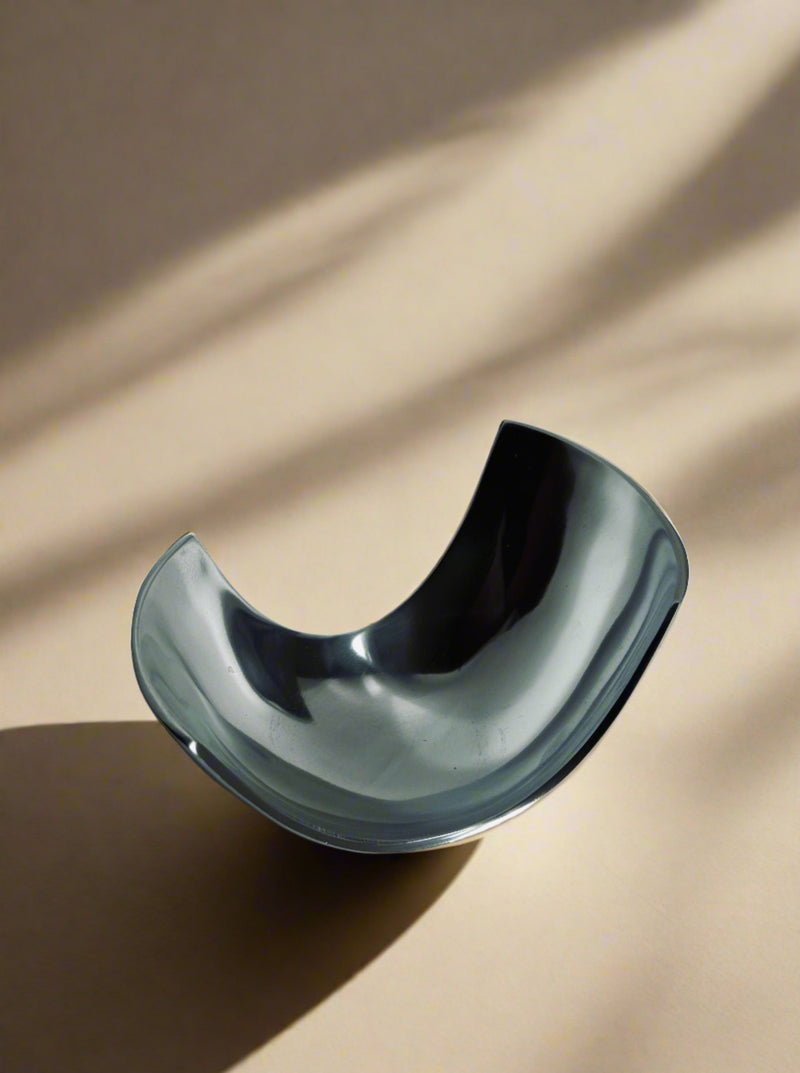 A sleek Modernist Decorative Dish by Flock Studio, with its unique curved shape, rests on a beige surface. Sunlight casts soft shadows across the background, accentuating the dish's shiny aluminium surface and elegant contours.