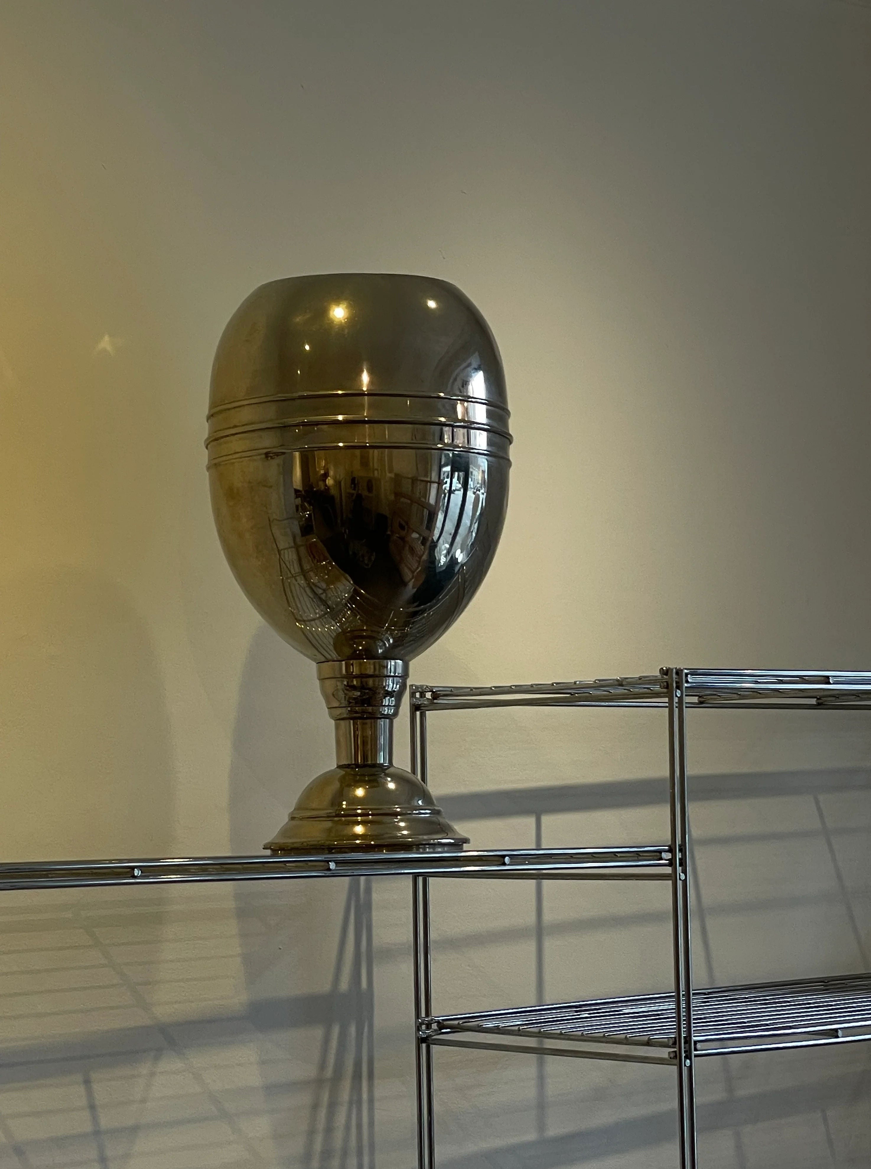 A large, shiny, steel trophy with a rounded cup and a slender base, sitting on a metal shelving unit against a plain wall.