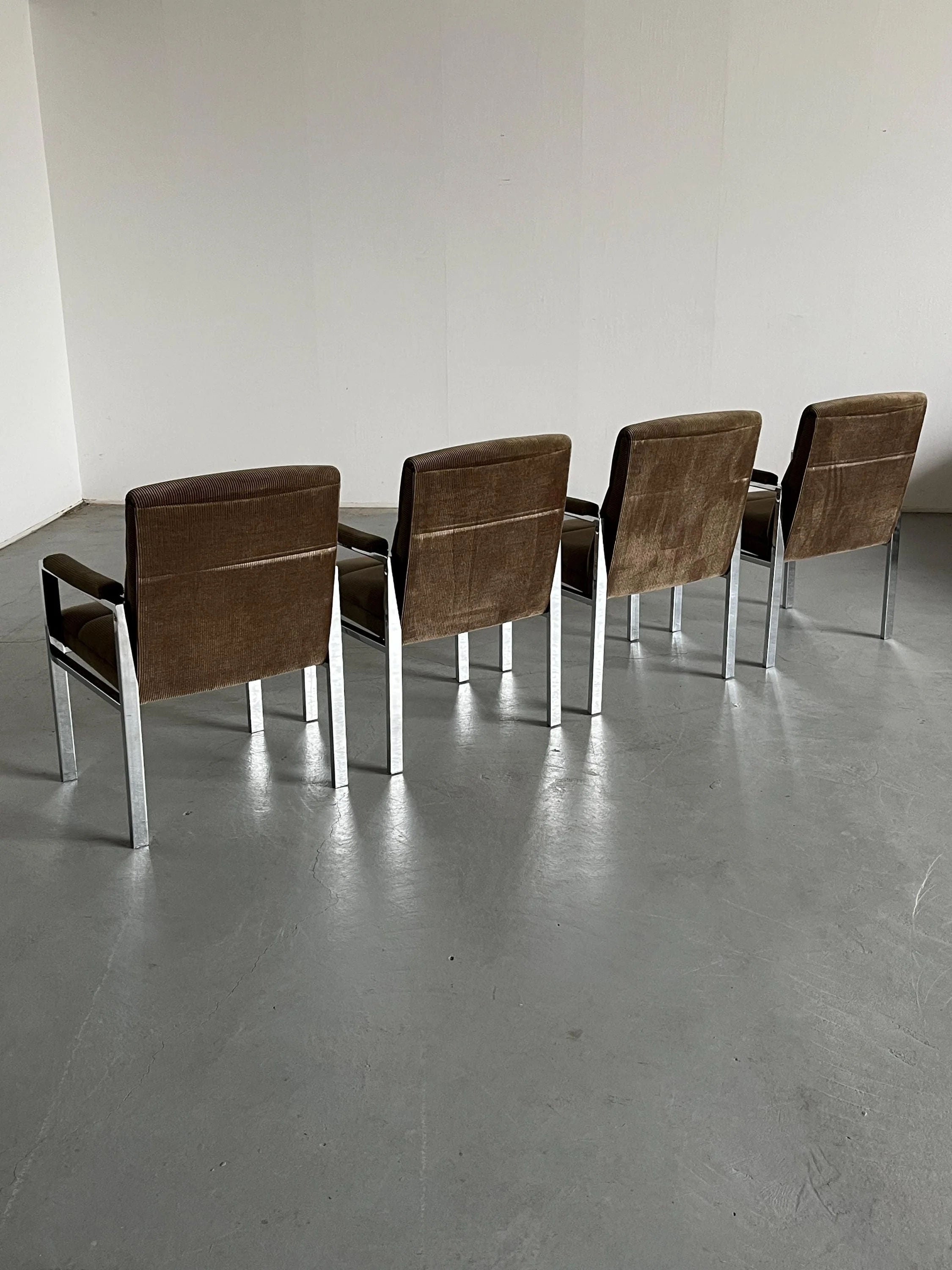 Four Cherry Cargo Mid-Century Armchairs from the 1970s, featuring brown corduroy upholstery and chromed steel frames, are aligned on a smooth, light gray floor against a plain white wall. Viewed from behind, their design reflects Vintage Mid-century modern aesthetics.