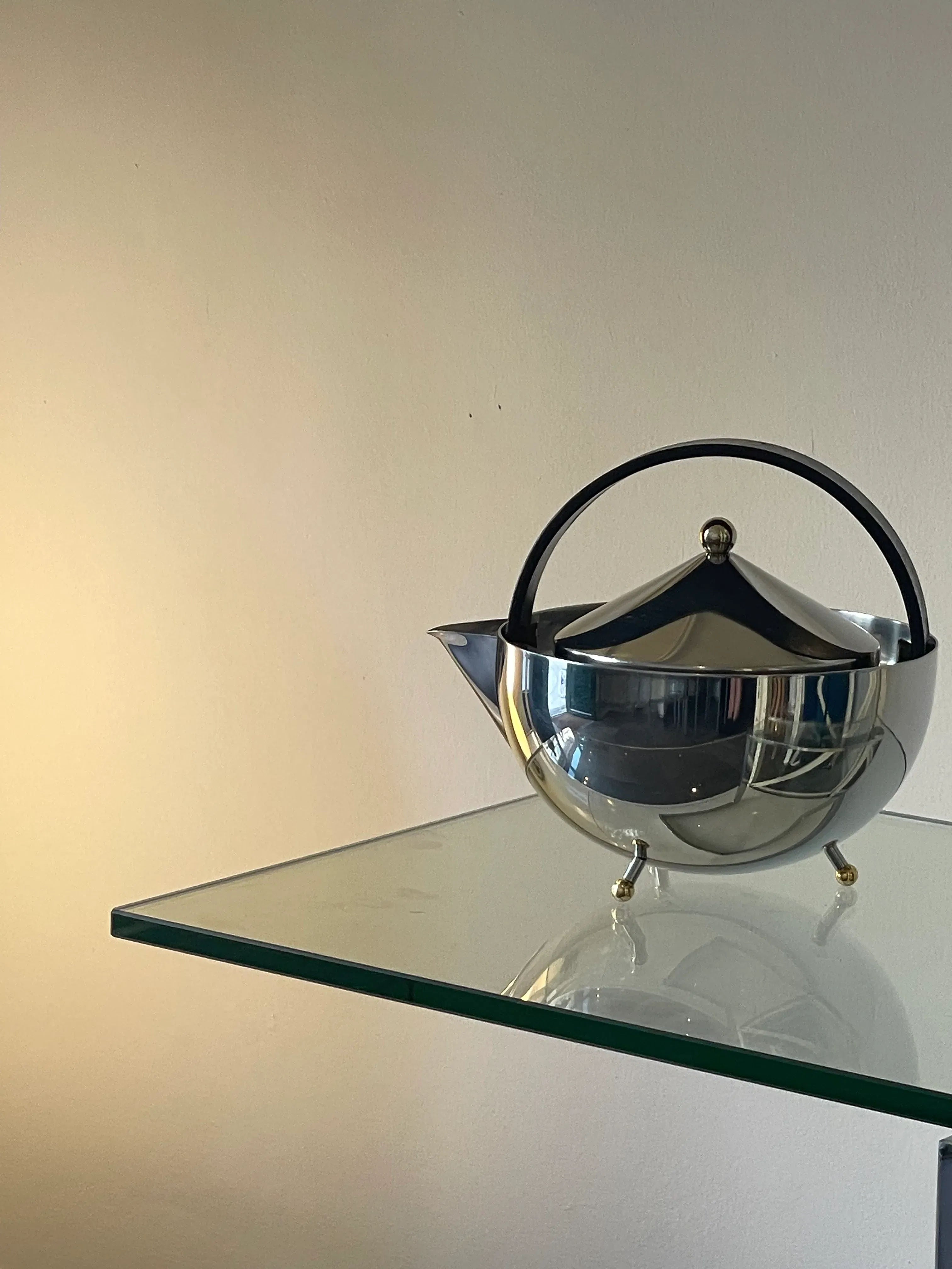 A sleek, silver Collect Cph Teaball Teapot with a black handle and lid is placed on the corner of a glass shelf. The teapot's polished surface reflects the surrounding light, highlighting its elegant design and built-in infuser basket. It is positioned against a plain, light-colored wall, creating a modern aesthetic.