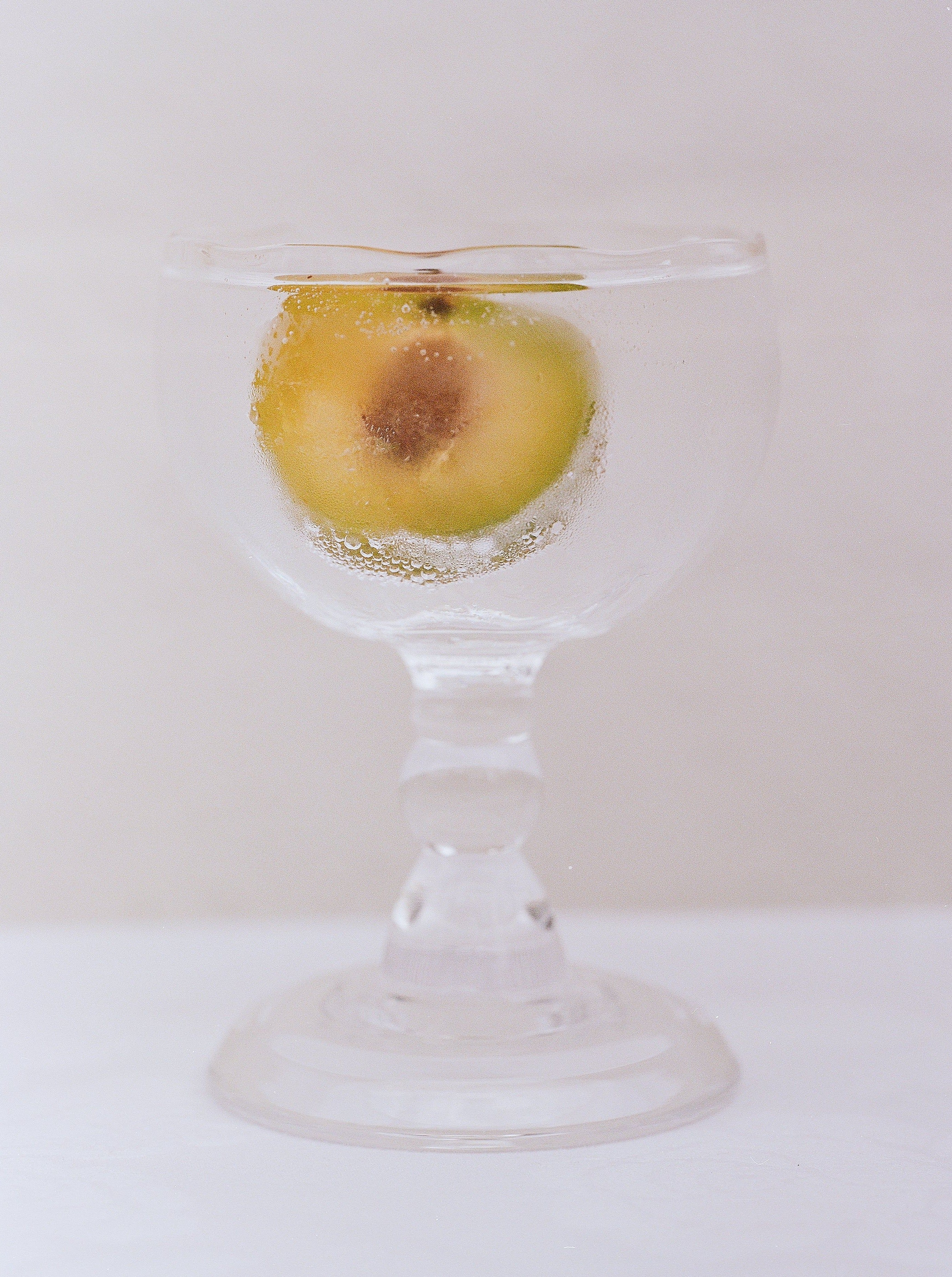 An apple submerged in water in a clear, mouth-blown glass goblet from the Akua Objects Alban Wine Glass Small collection, with bubbles clinging to its surface, placed against a soft beige background.