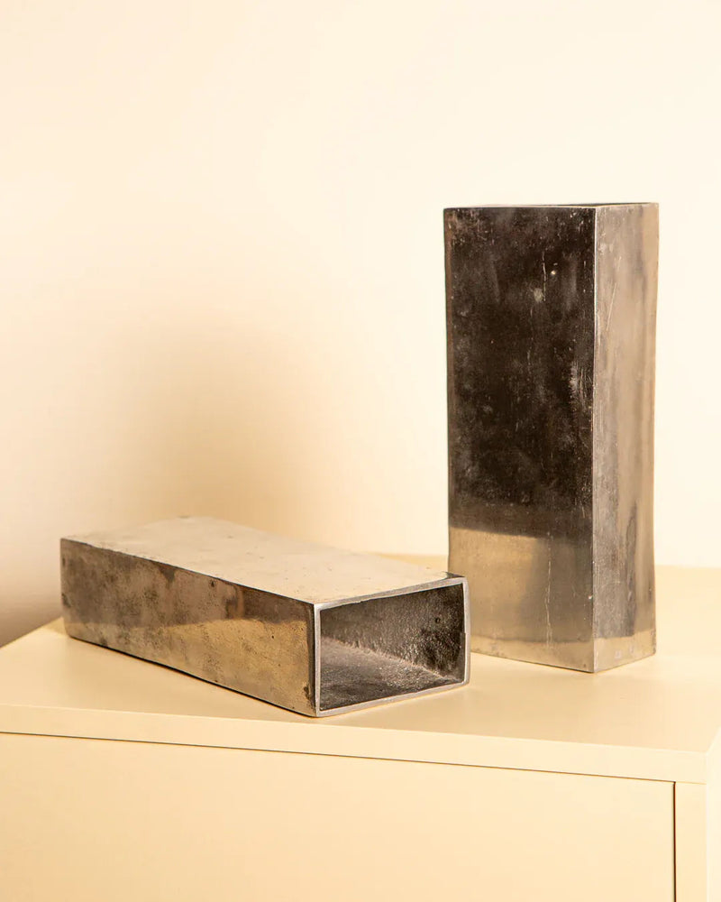 A pair of 80's Italian aluminum vases by Treaptyque, one upright and one lying down, are set on a light surface with a cream backdrop. Their reflective finish and varied textures evoke vintage design.