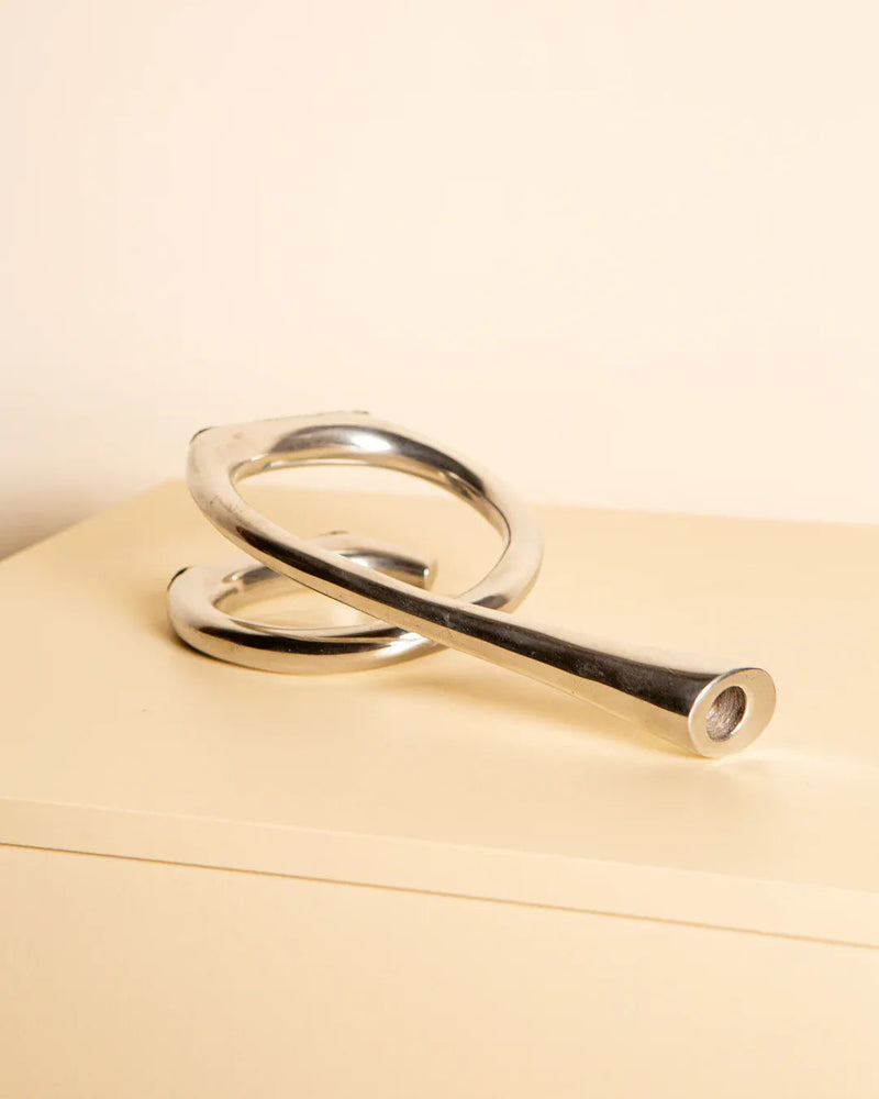 The Cast Aluminum Candleholder 80's from Treaptyque is a sleek, modern, silver abstract sculpture with smooth curved lines embodying minimalist design, elegantly displayed on a beige surface against a light background.