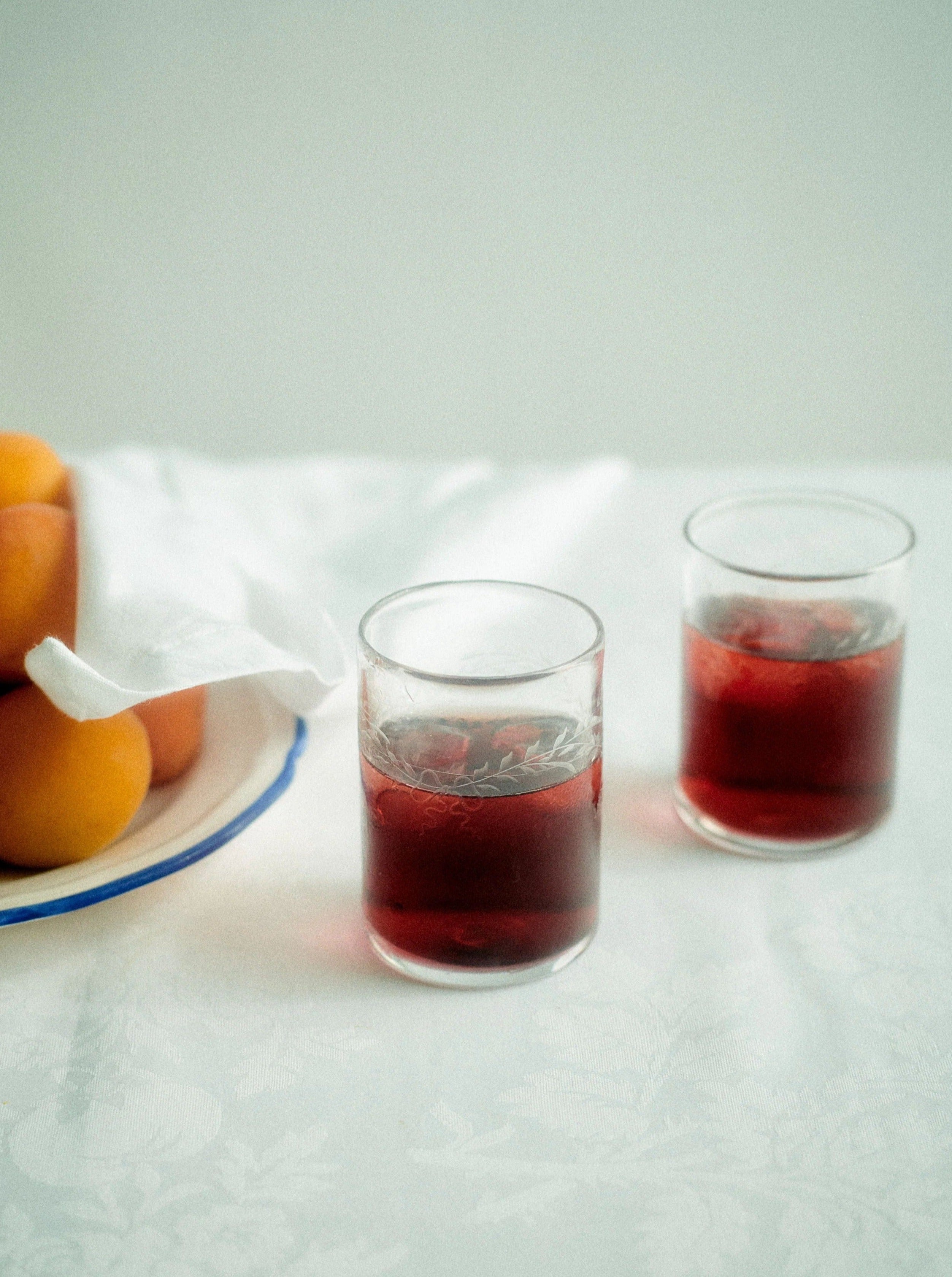 Two glasses of red drink in Barbro Water Glass - Set of four by Akua Objects on a lace-covered table with a bowl of oranges in the background, creating a serene and inviting scene.