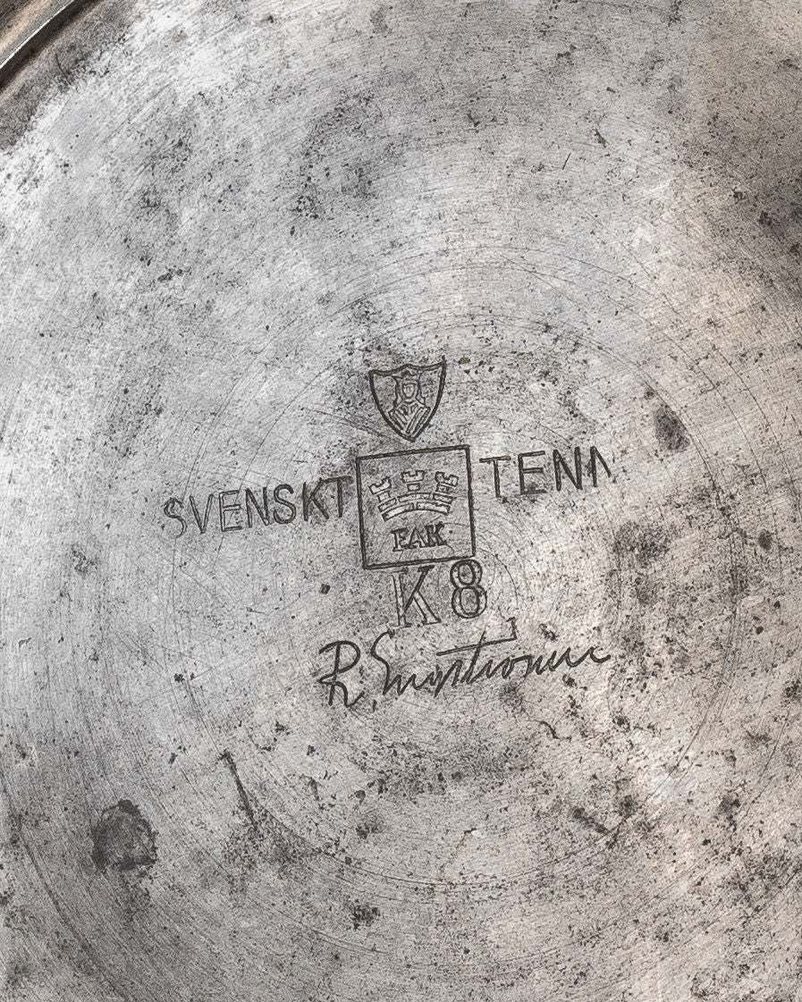 A close-up of the Large Modernist Pewter Amphora by Spigel from the 1930s reveals an engraved metal surface with a coat of arms featuring three crowns, the words "SVENSKT TENN," "FALK," and "K8," plus a signature resembling "R. Sundström." The Scandinavian piece has a worn, tarnished look.