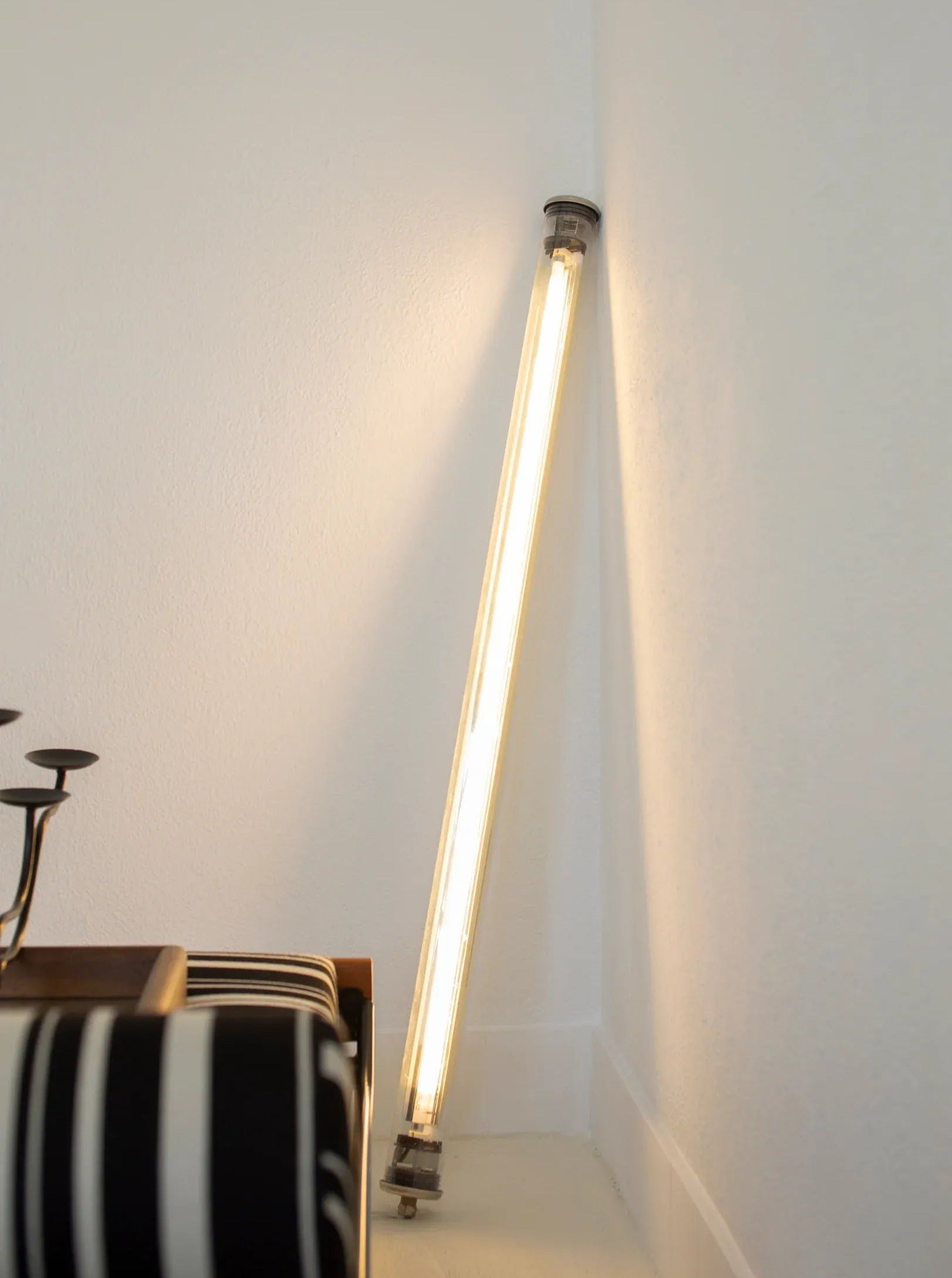 A CU-RA-TED Sammode Light Tube is propped against the corner where two white walls meet. Below it, part of a piece of furniture with black and white striped upholstery is visible in the bottom left corner, showcasing French architectural lighting elegance.