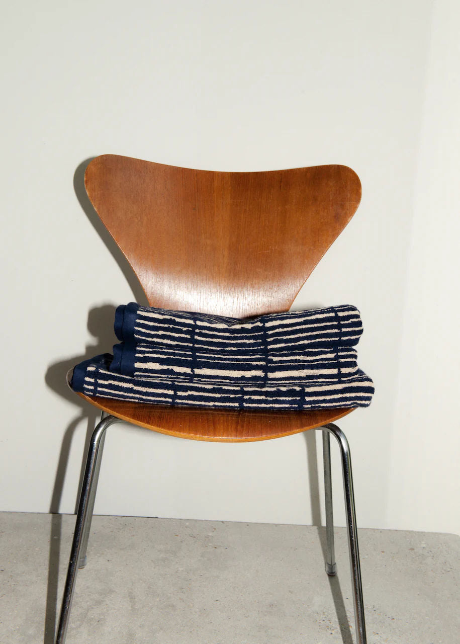 A sleek wooden chair with metal legs holds a folded, patterned Ester Bath Towel. The 100% cotton towel features a checkerboard-style design in blue and white, exuding luxurious comfort. The chair is placed against a plain white wall, and the floor is a light grey color.