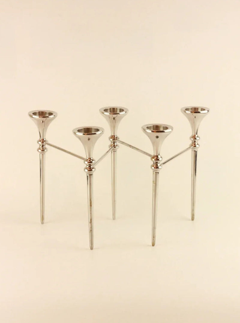 The Scandinavian Modular Candelabra by Boga Avante Shop is a modern, metallic candelabra with five candle holders arranged in a symmetrical design. The holders are elevated on slender, modular arms connected to a central frame, which has three legs for support. The overall aesthetic is sleek and contemporary, reminiscent of a vintage chrome chandelier.
