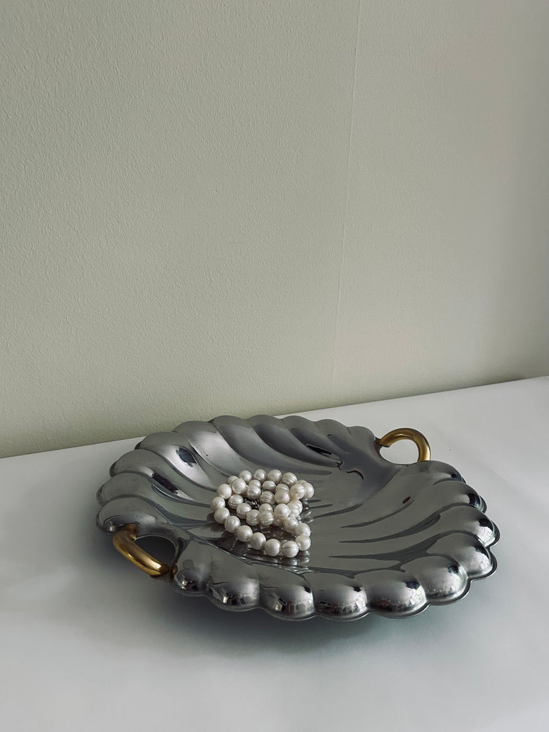 Crafted in Italy, the Flock Studio Italian Stainless Steel Shell Tray features a luxurious design with scalloped edges and two gold handles. It elegantly showcases a string of pearls arranged in a heart shape, resting on a white surface against a plain beige wall background.