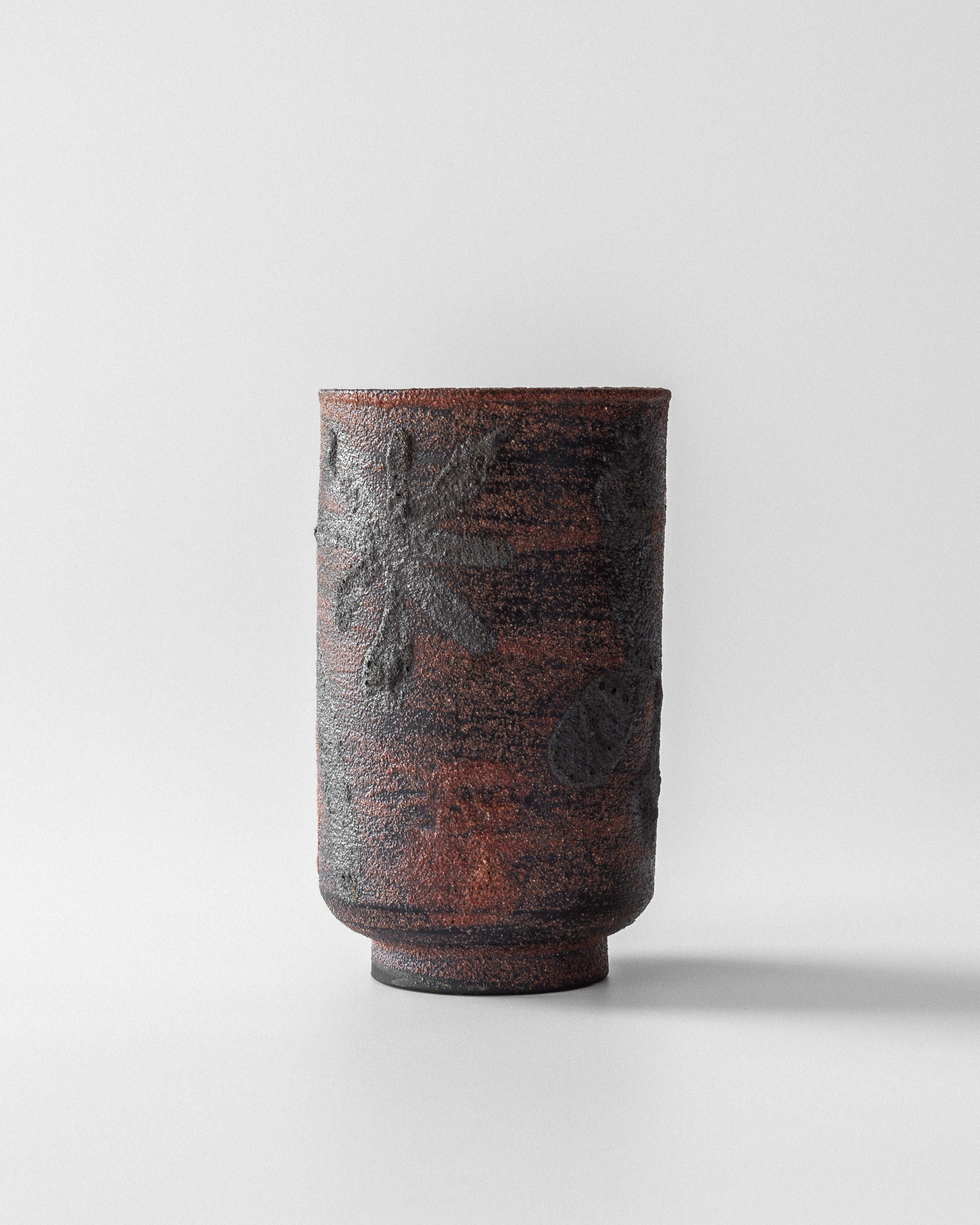 The Modernist Vase by Jaap Dommisse for Spigel is a textured, cylindrical piece in dark earthy tones with subtle raised floral patterns on a plain white ceramic background.