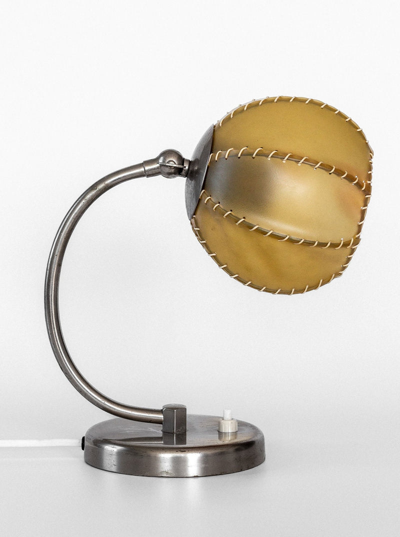 The Spigel 1930s Art Deco Metal Lamp features a spherical, yellowish lampshade with stitched seams, mounted on a curved chrome body and a round base. The base includes a switch and a white power cord. Its design captures the mid-20th century aesthetic perfectly, making it a true collectible.