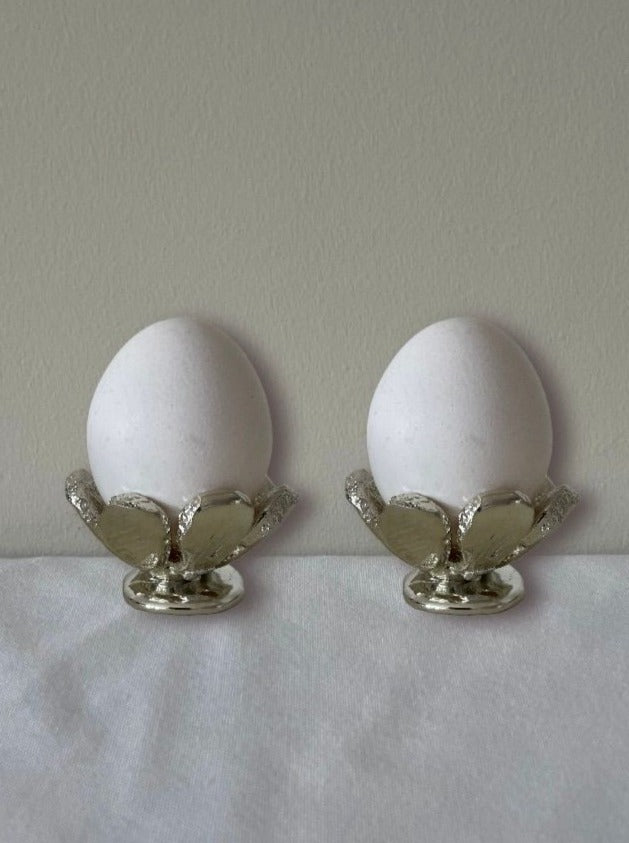 Two white eggs stand upright in The Tin Collection / FLWR egg cups by Studio Formbart, crafted from tin in a leaf shape, displayed on a white surface with a light background.
