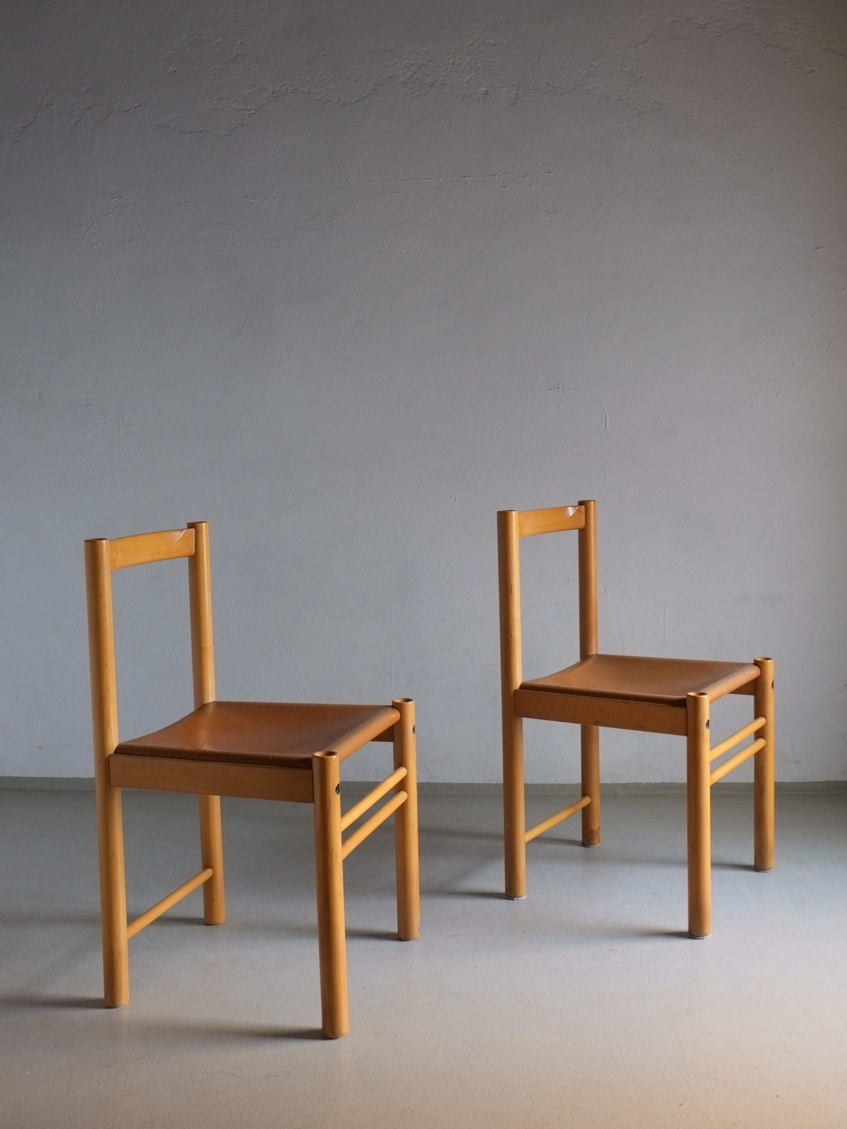 Two of the 6 Beech Wood Cognac Leather Chairs from Ibisco, Italy, crafted in the 1960s and sourced from the Veter Vintage brand, are placed on a smooth, light-colored floor. The setting is against a plain, neutral-toned wall, creating a simple and elegant composition.