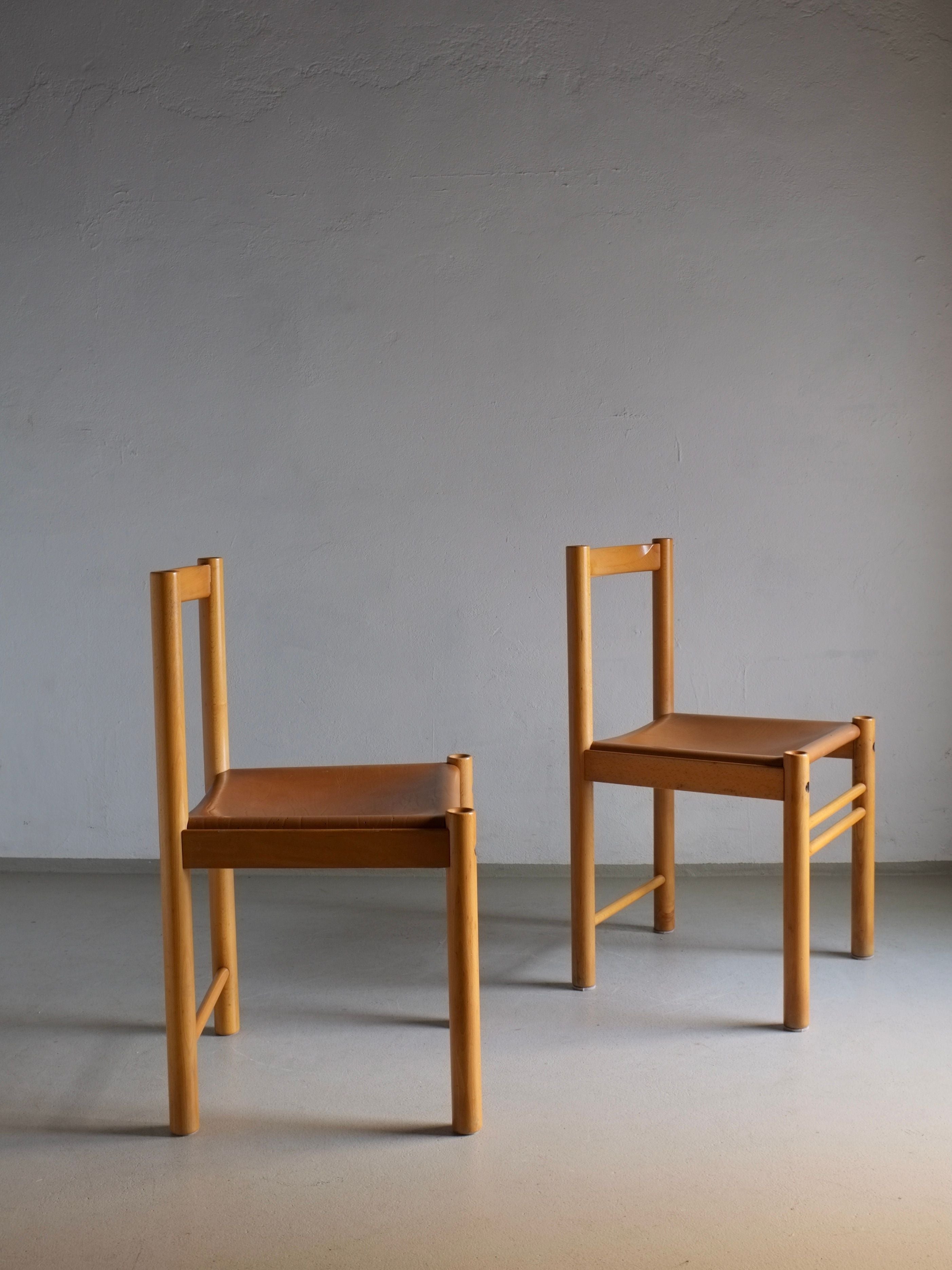 Two Veter Vintage Ibisco dining chairs from the 1960s, crafted from beech wood and featuring cognac leather seats, rest on a light gray floor against a plain gray wall. Positioned at slight angles to one another, they create a simple and modern composition while evoking vintage charm.