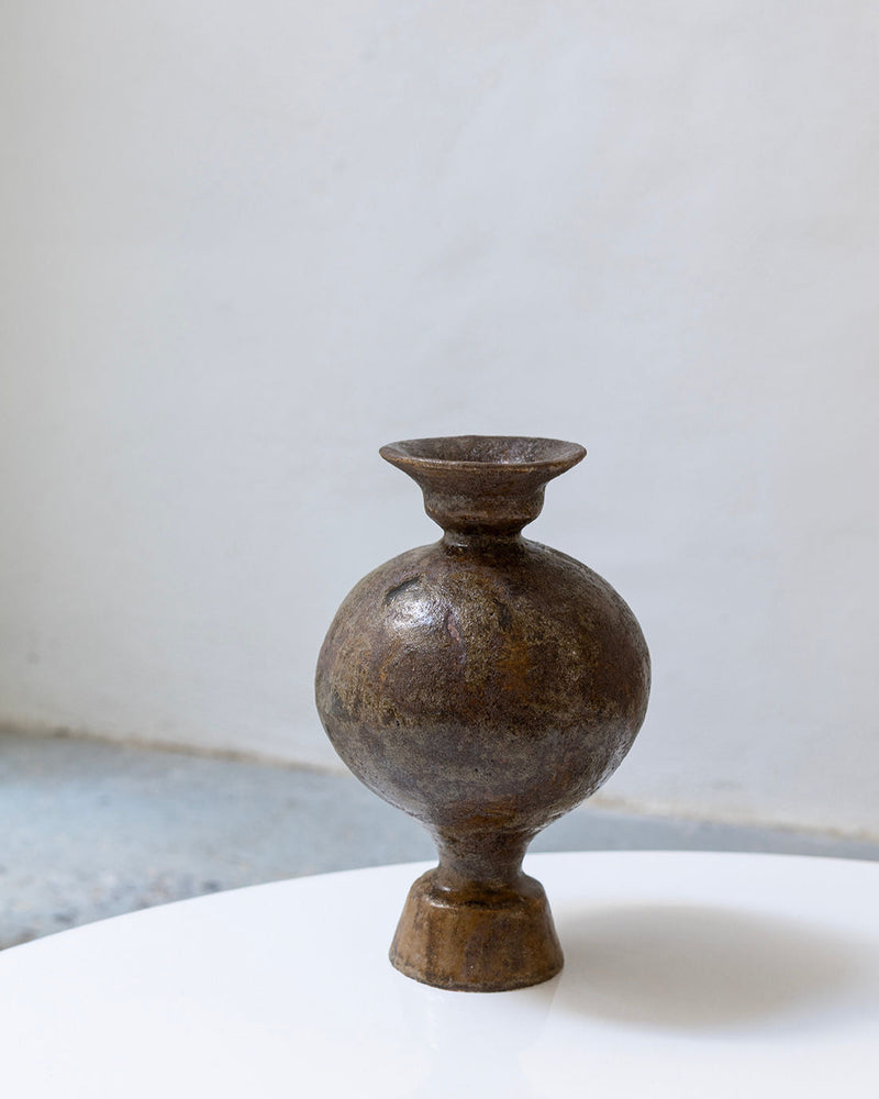 The Lekytho Bufalo Vase by Canoa Lab, a round rustic brown stoneware piece with a narrow neck and base, sits on a white surface against a light, minimalist background.