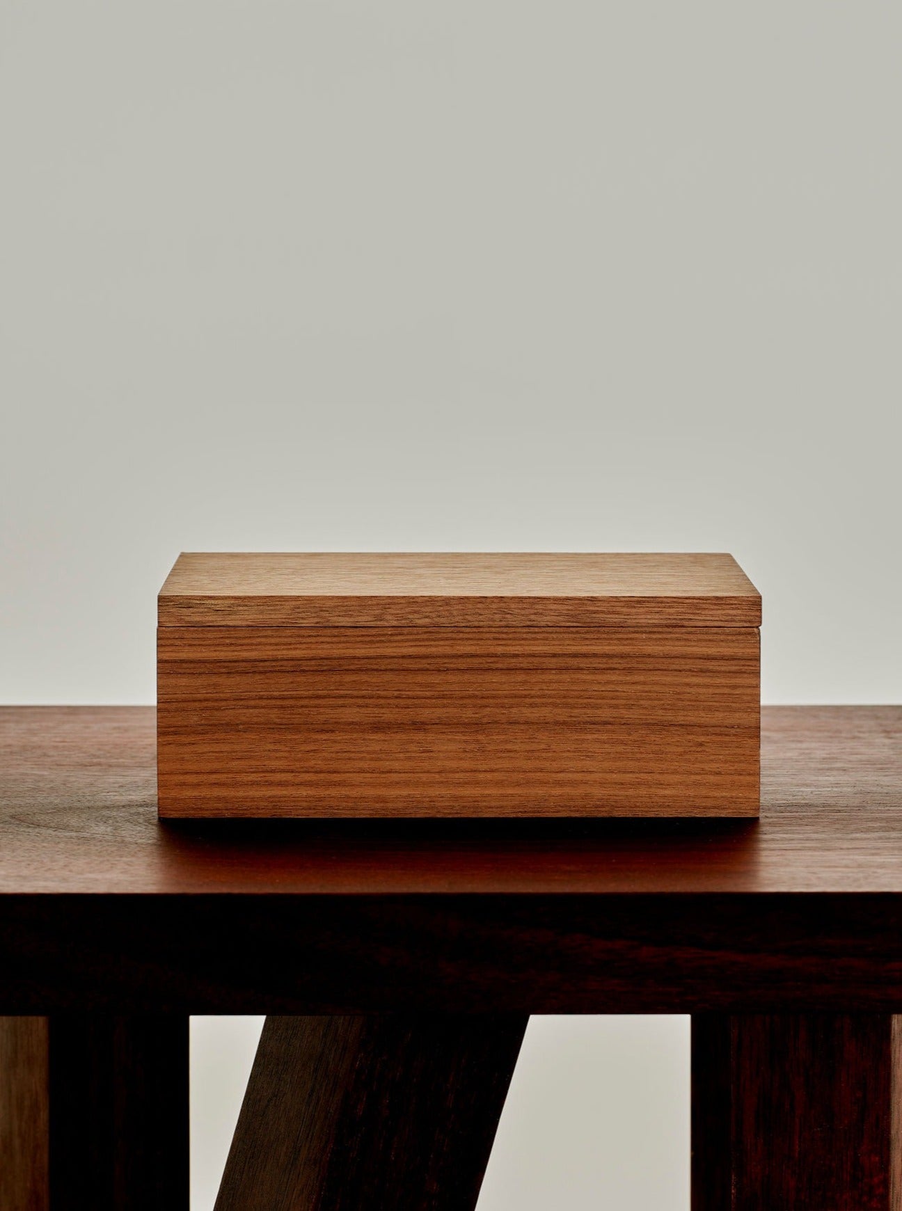 A small, rectangular Miter Box by MOCK STUDIO sits centered on a larger, dark wooden table with an abstract leg design, both against a plain, light-colored background. The box and table have distinct wood grain textures and minimalist designs, highlighting their high grade material.