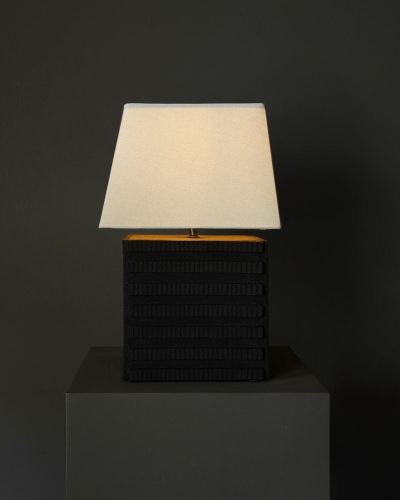 The Akune Ceramic Lamp - Number One by Studio OSKLO, featuring a textured black square base and rectangular beige shade inspired by post-Bauhaus brutalism, is illuminated on a dark surface against a plain dark gray background.