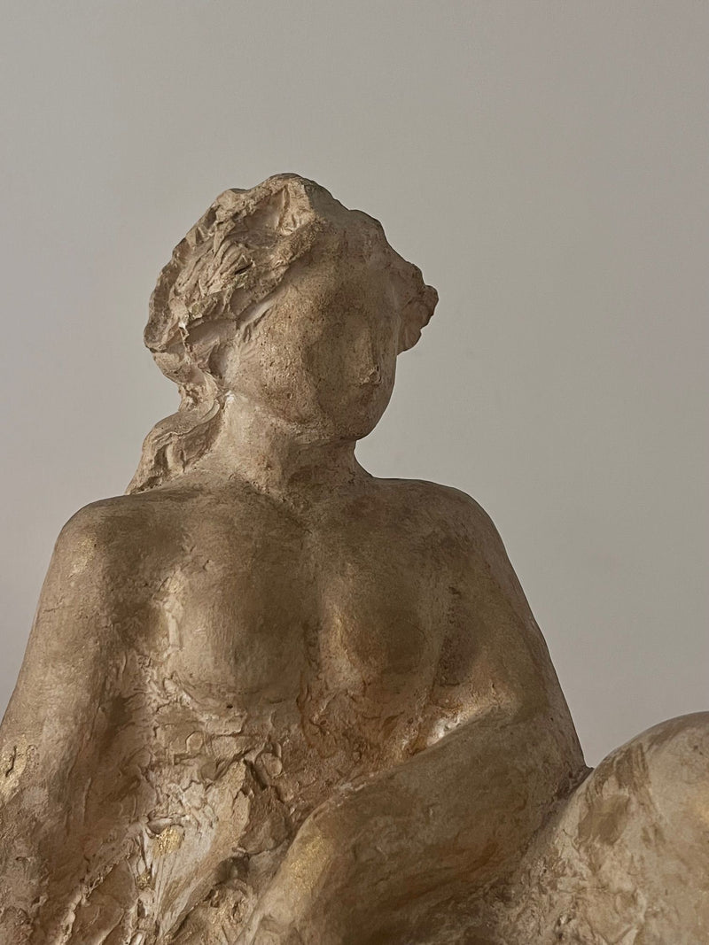 A close-up of Sculpture by Sakari Tohka 1938 by Médecine—a textured beige plaster figure of a seated woman with wavy hair and minimal facial features, set against a light, plain background.