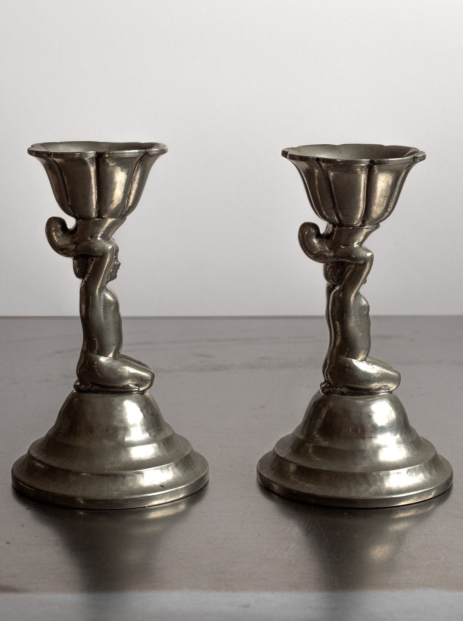 Two silver-hued metal candle holders from the 1930s Art Deco era by Spigel, Denmark. Each holder, reminiscent of antique pewter candlesticks, features a human figure in a kneeling position, supporting the candle platform above their heads. The bases are round and tiered, enhancing the elegance of the neoclassical design.