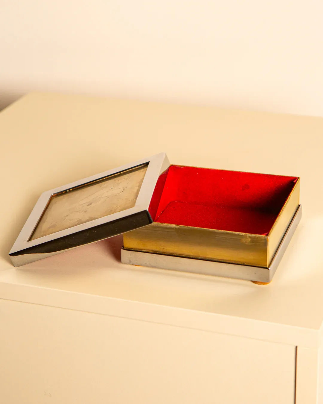 An Italian Treaptyque box from the 60s, with a gold exterior and red interior, sits on a light surface. Its slightly open lid showcases a reflective silver-plated frame.