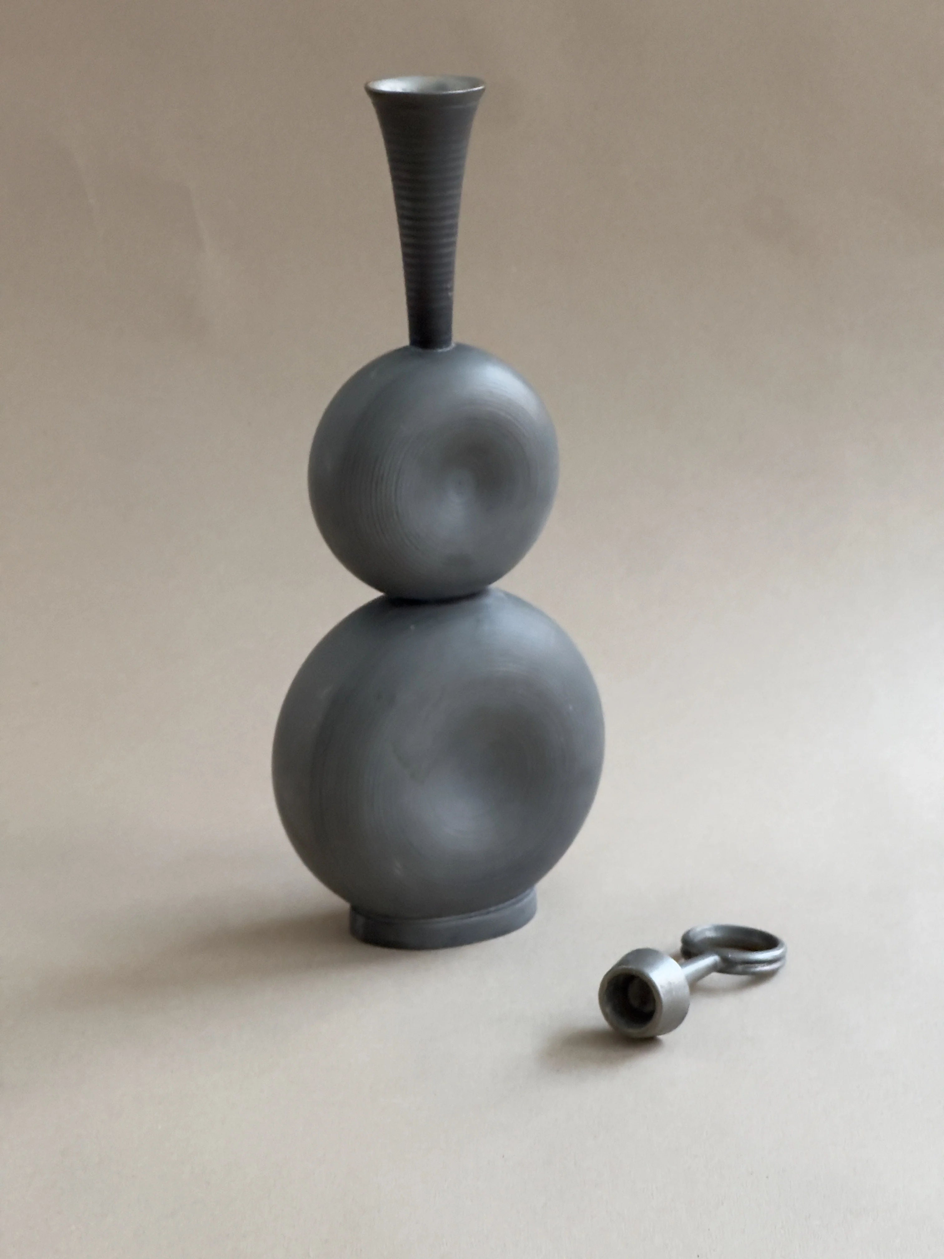 On a neutral background, a modern abstract gray sculpture by Gunnar Havstad with a circular base and tall, narrow top stands beside the Modernist Pewter Decanter from Galerie Storm, reflecting similar material and style with its round shape and attached ring.