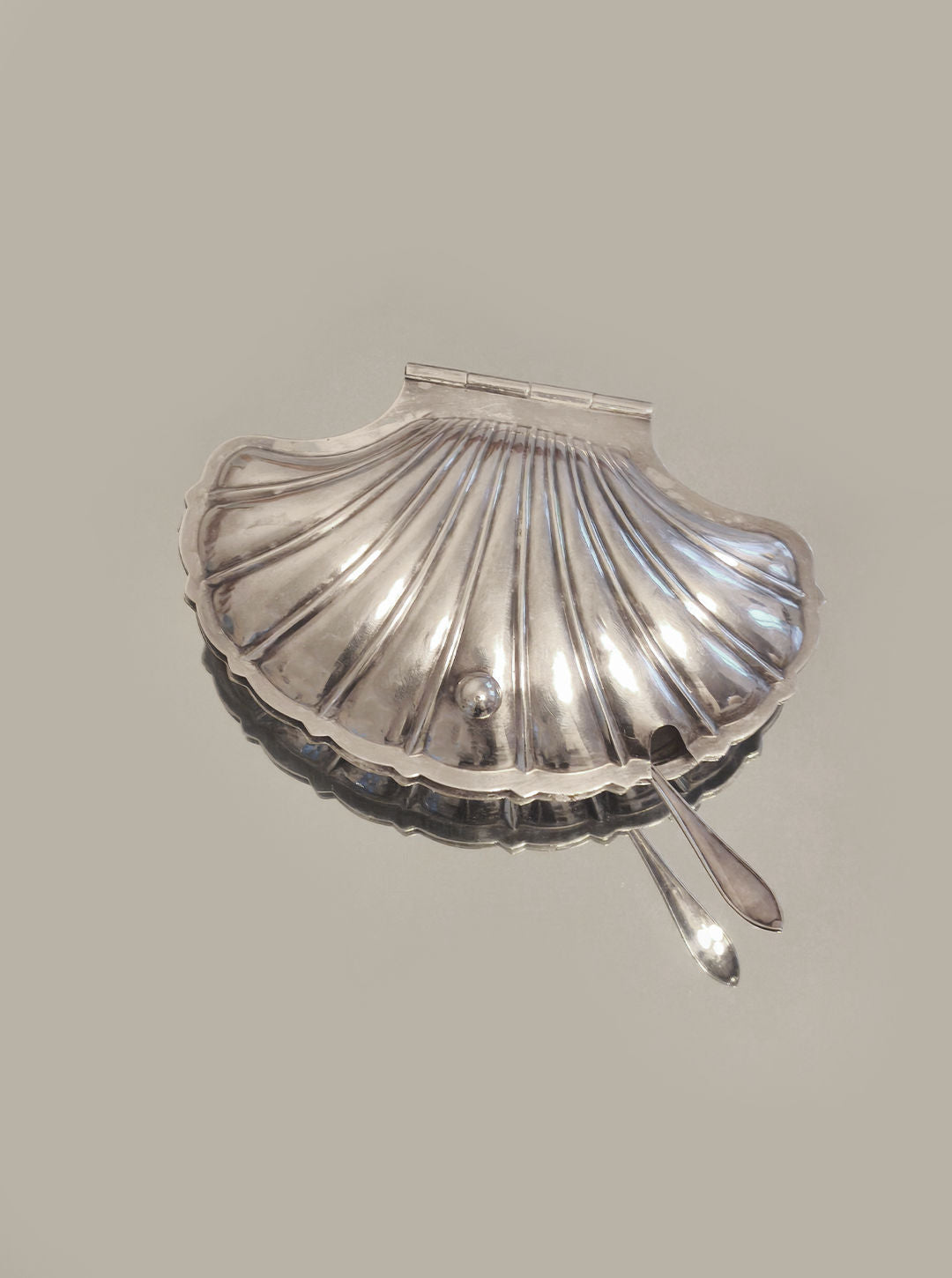 The Shell Butter Dish by Les Objoies is a silver-plated, scallop shell-shaped butter dish complete with a small spoon, ideal for elegant dining settings and set against a plain background.