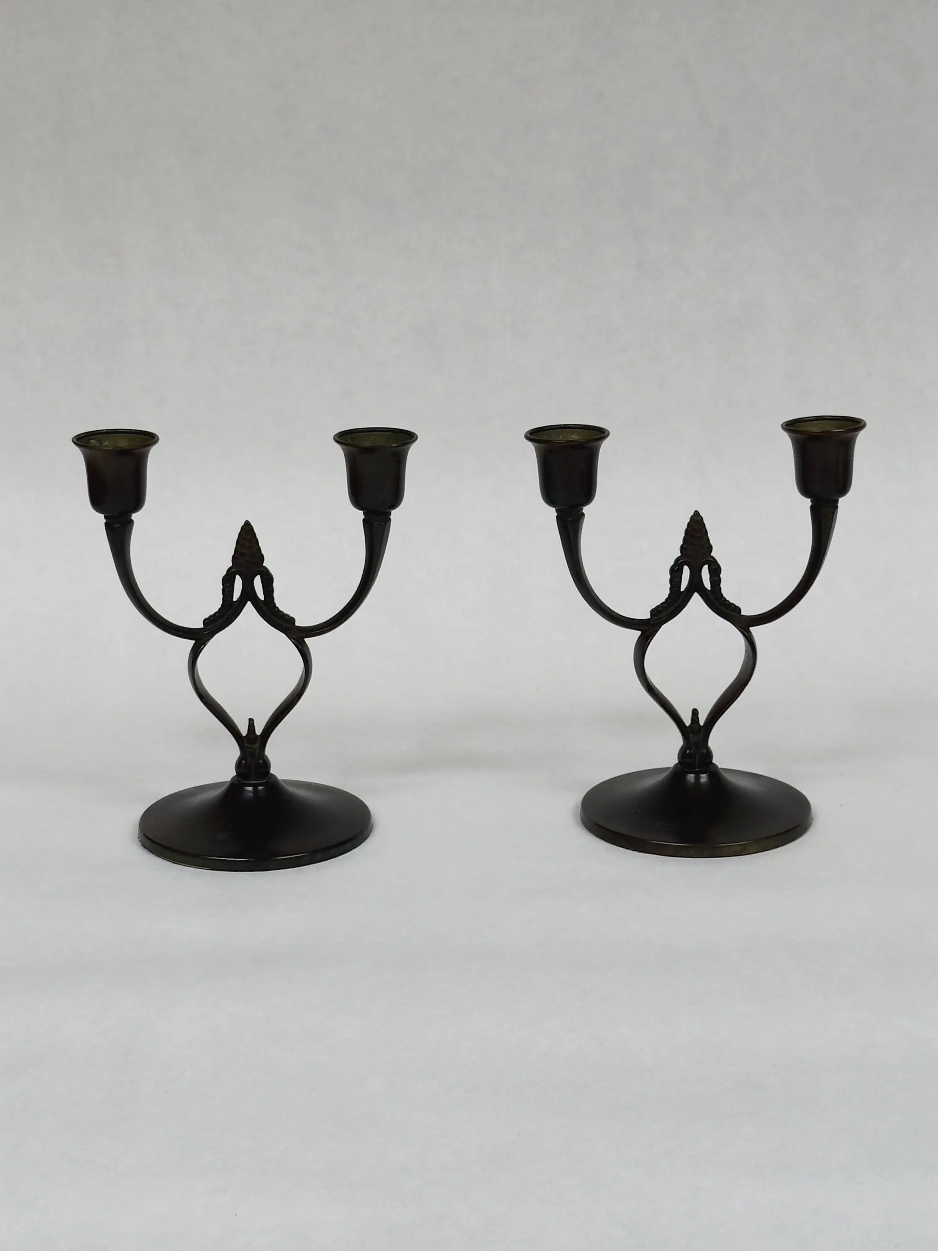 Two Galerie Storm antique bronze candlesticks with an elegant twist design hold two candles each, reminiscent of Art Nouveau style. They are symmetrically placed on a light gray surface against a matching background.