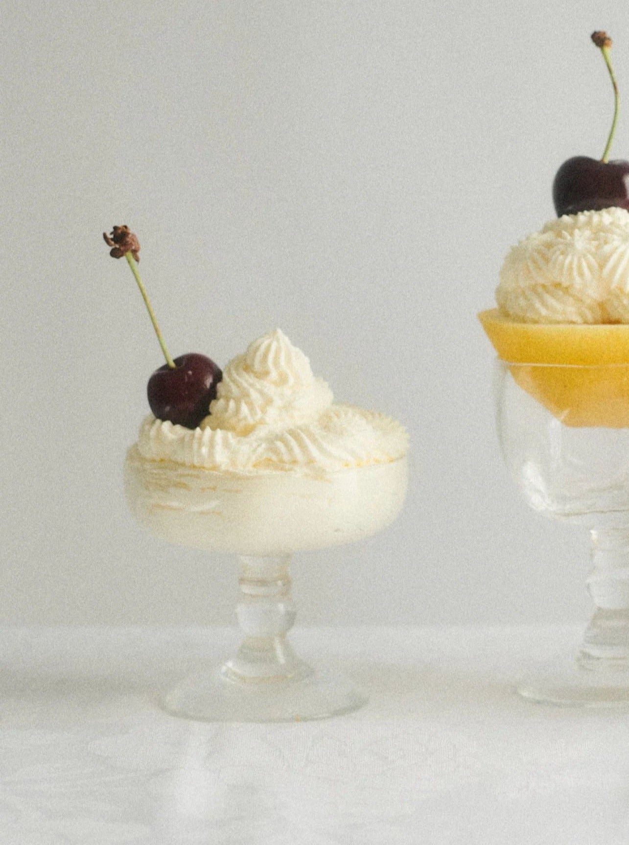 Two elegant Akua Objects Alban Champagne Glass dessert glasses filled with whipped cream and slices of mango, garnished with fresh cherries on top, set against a soft, light grey background.